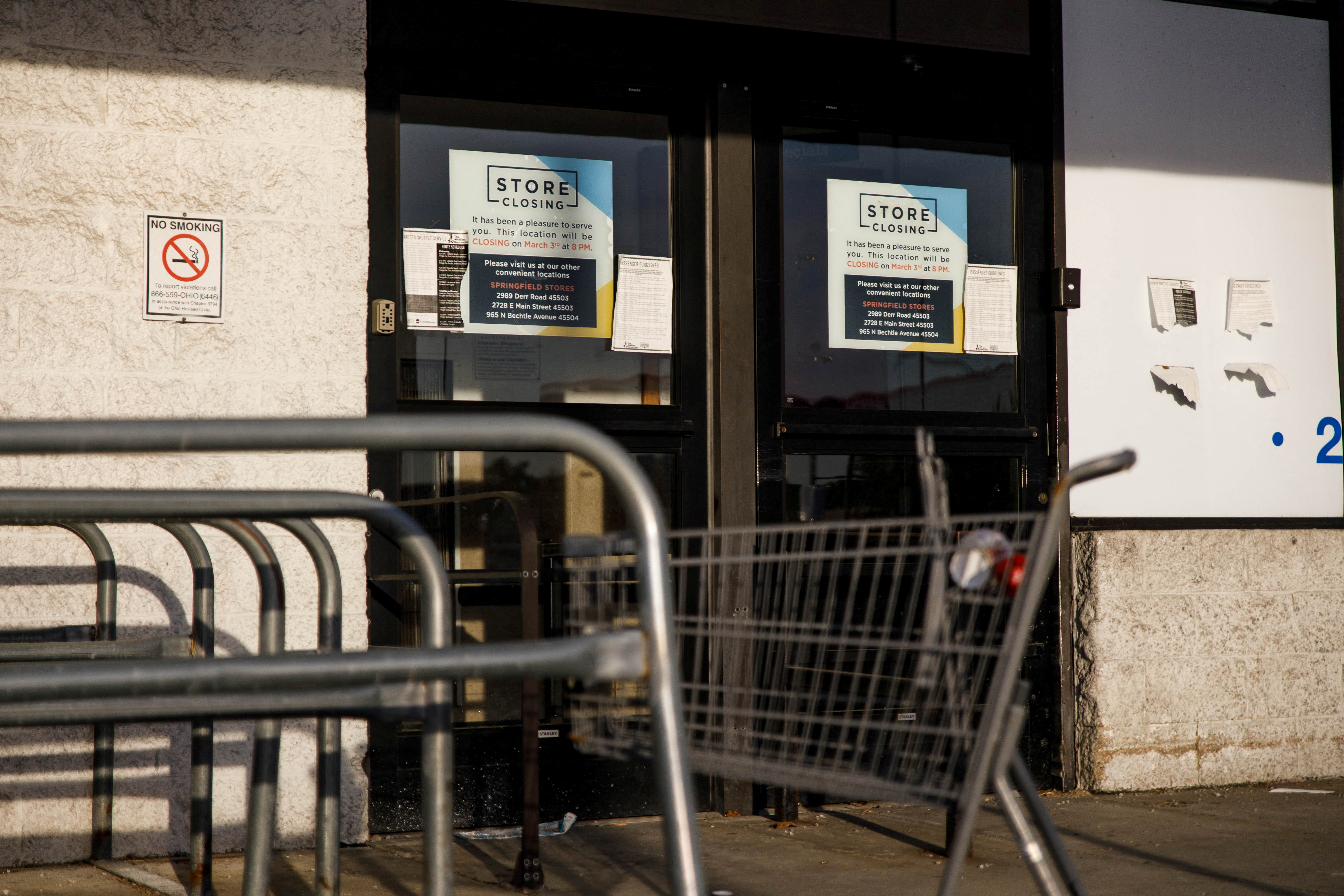 An empty shopping cart sits in front of the double doors of a super market, which bears the sign "Store Closing"
