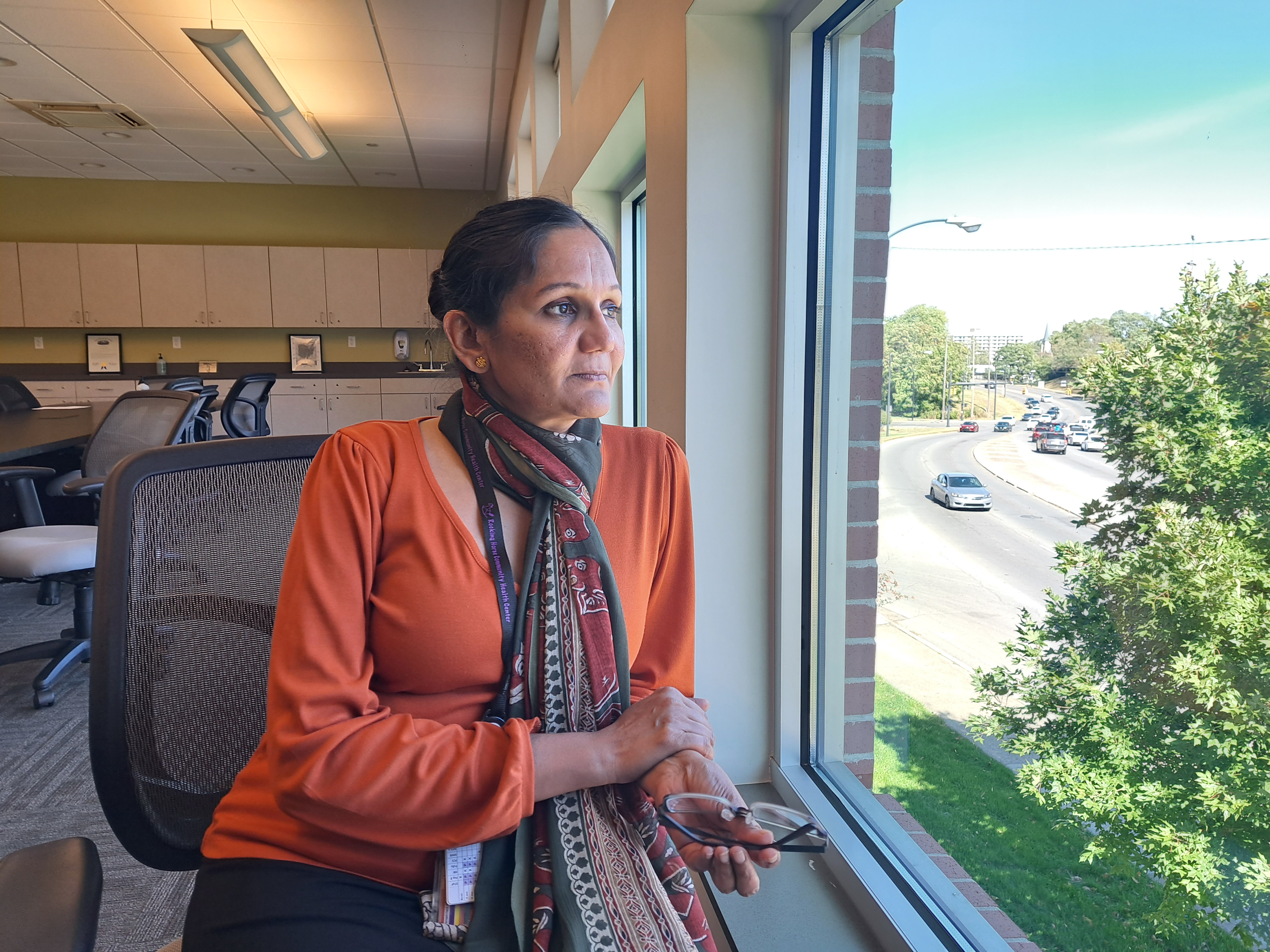 A woman in an orange, long-sleeved shirt and scarf leans against a window in the upper story of an office building, looking out toward the street below.