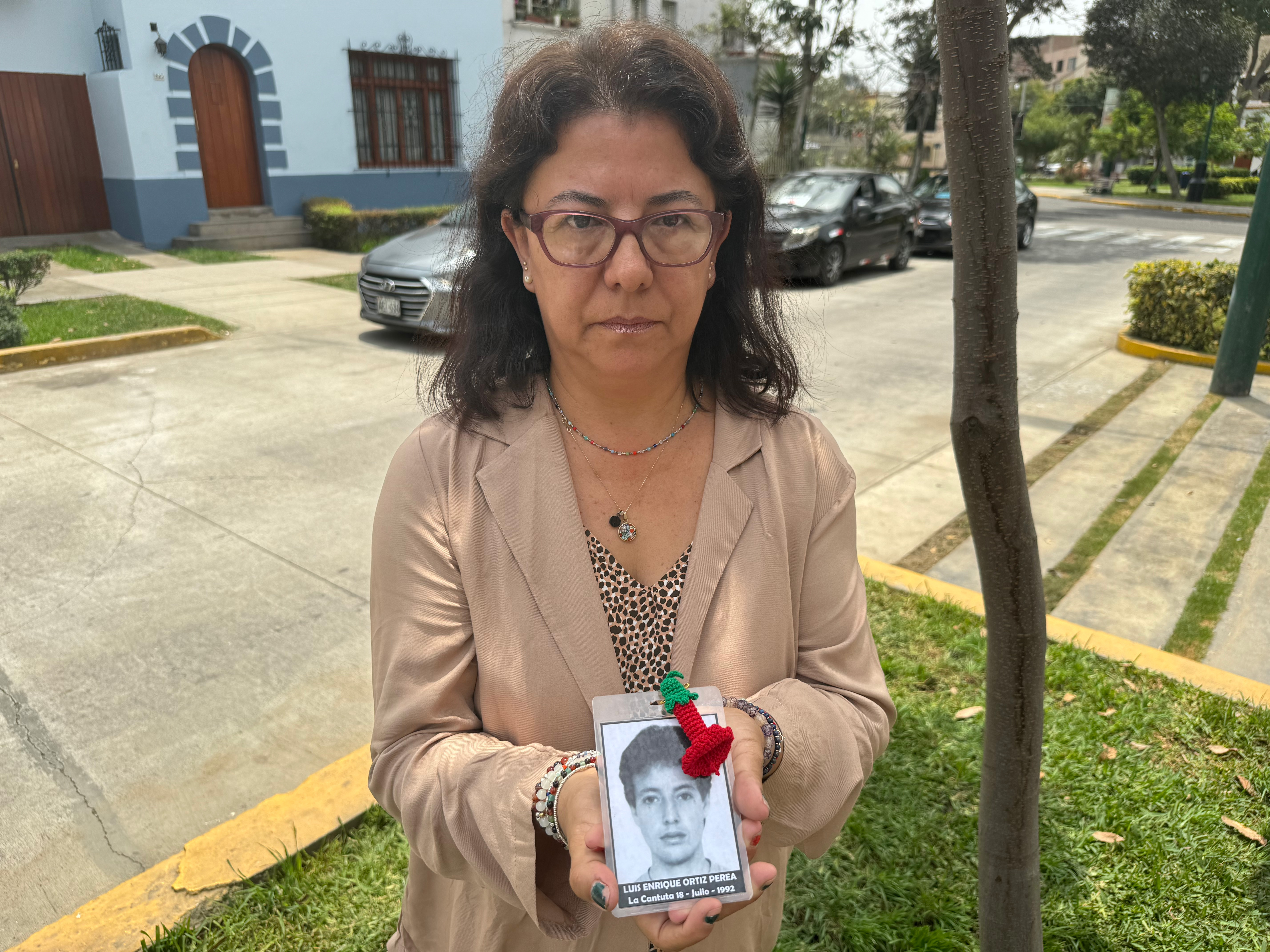 A woman in a tan blazer and glasses holds up a small black-and-white photo of a young man, with his name and the date he disappeared.