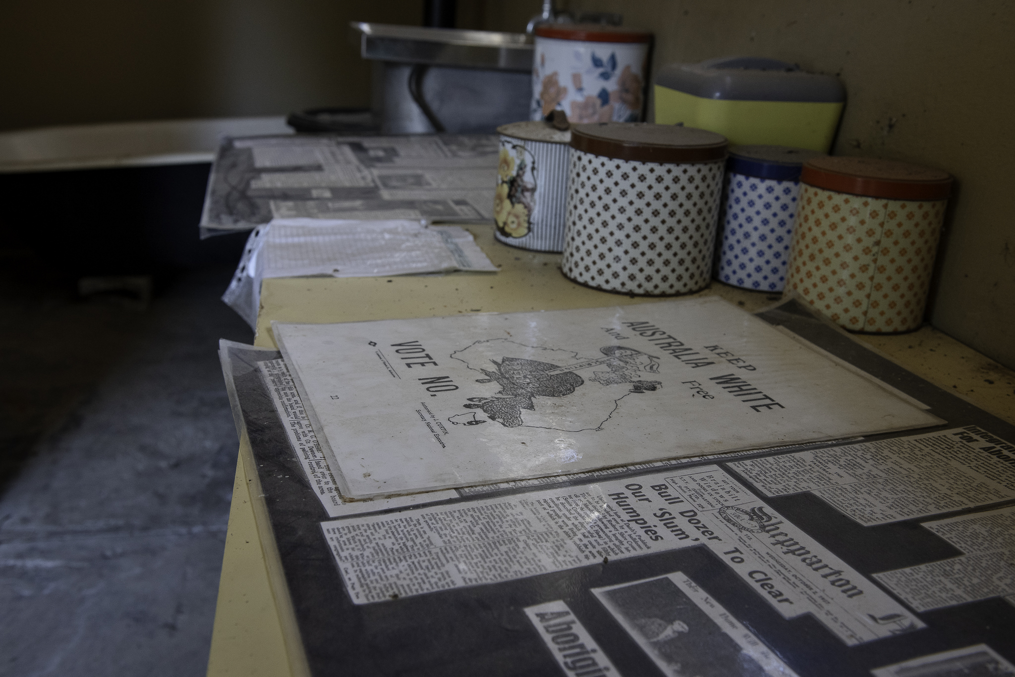An old leaflet saying 'Vote No to Keep Australia White' on a table in a replica of a house built for Aboriginal people. There are tins on the table as well as other documents.