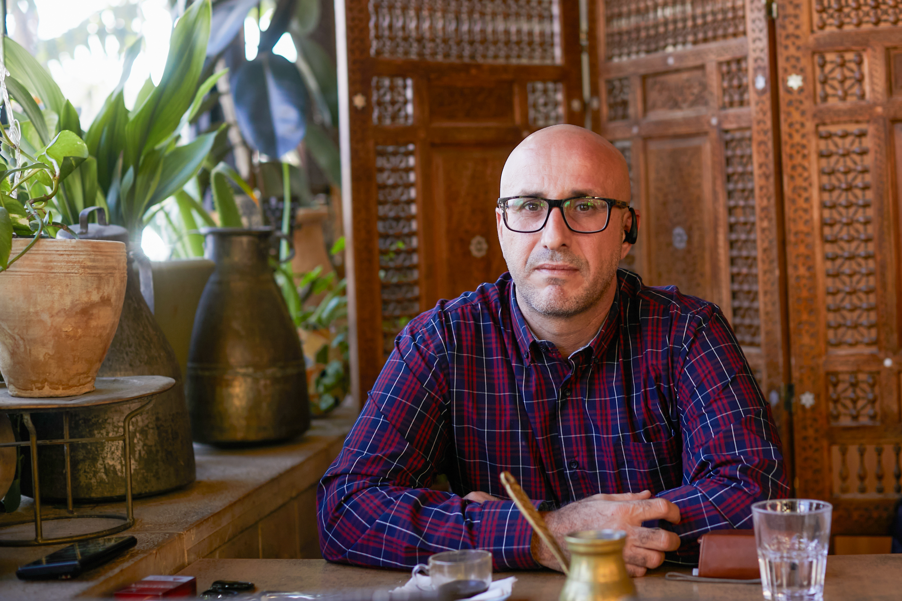 Shadi in a plaid shirt seated at a cafe table with coffee in front of him