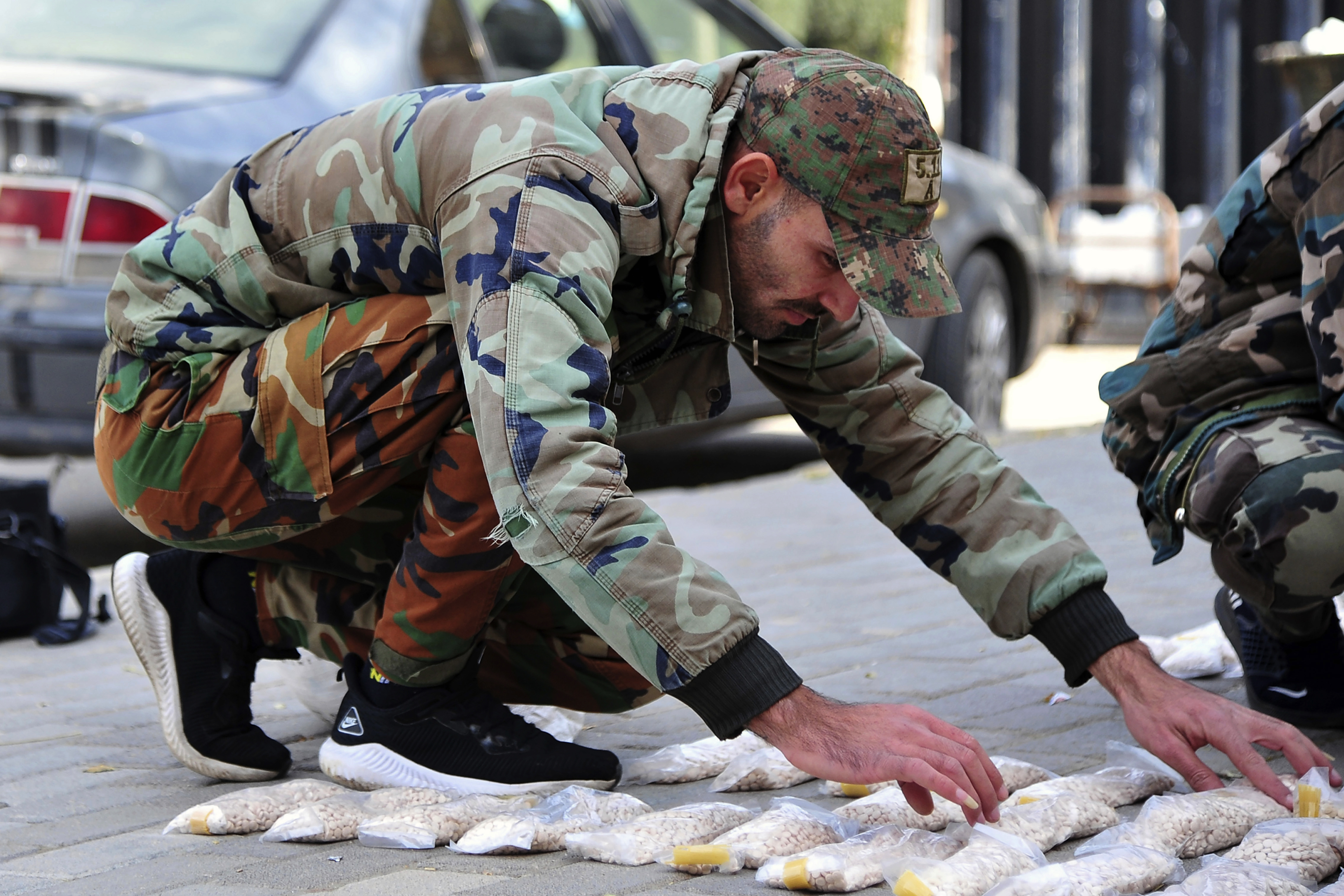A Syrian soldier kneels over rows of captagon in little packets, arranged on the pavement.