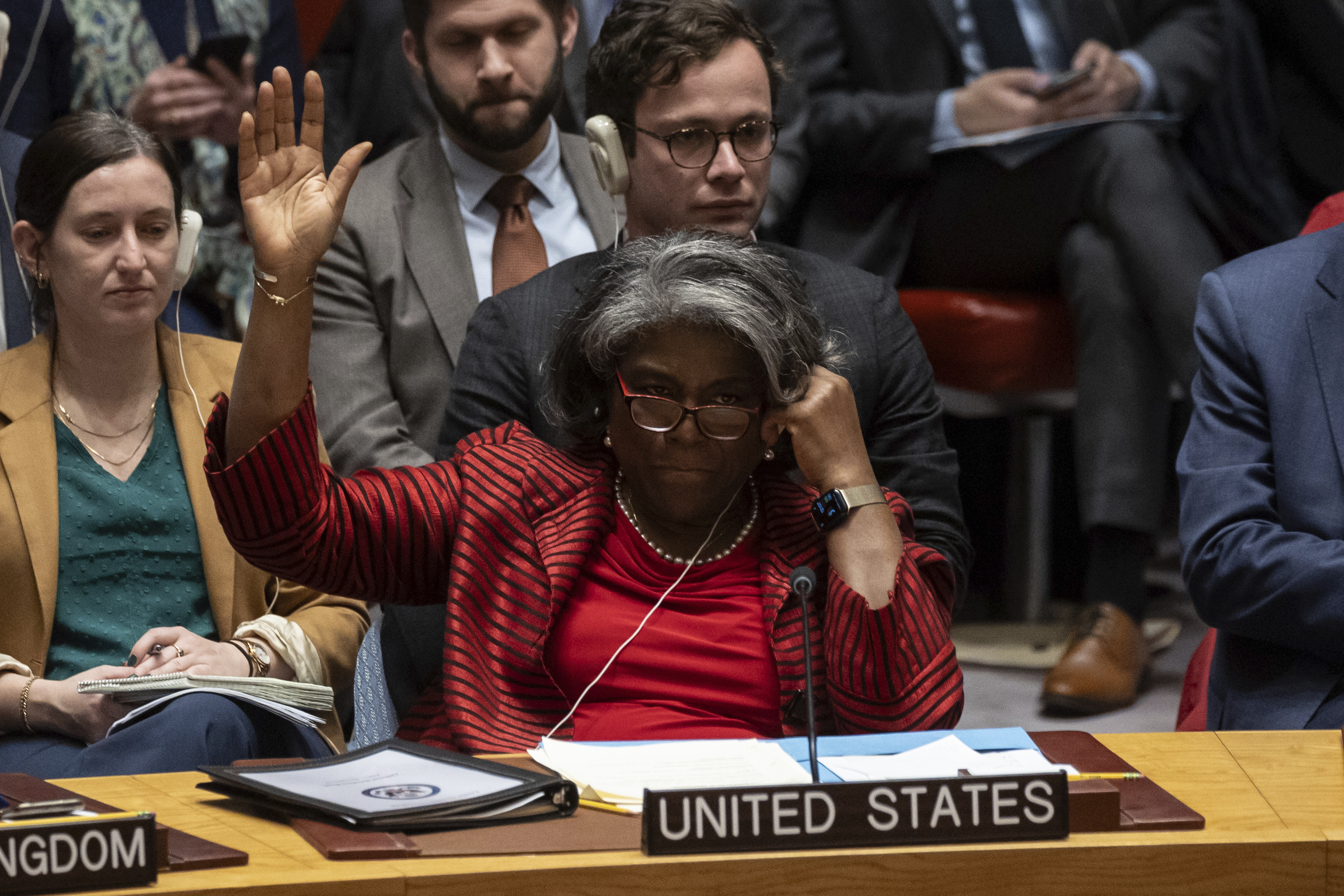 A woman wearing a red suit and red glasses raises her hand to vote behind a plaque saying United States