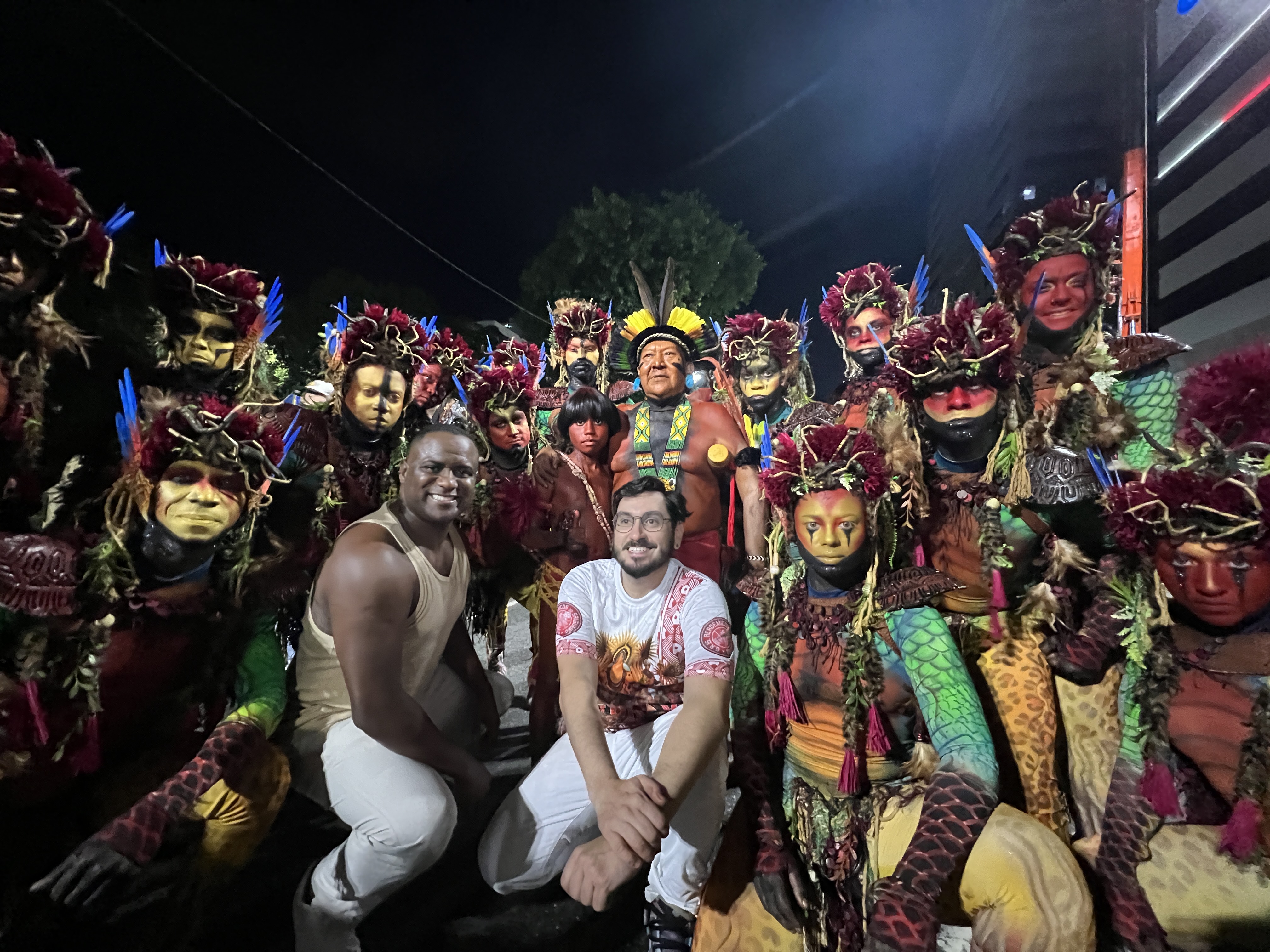 Davi Kopenawa, wearing a headdress of yellow and green feathers, stands in the centre of a group of Carnival parade participants, dressed in body paint and elaborate headdresses.