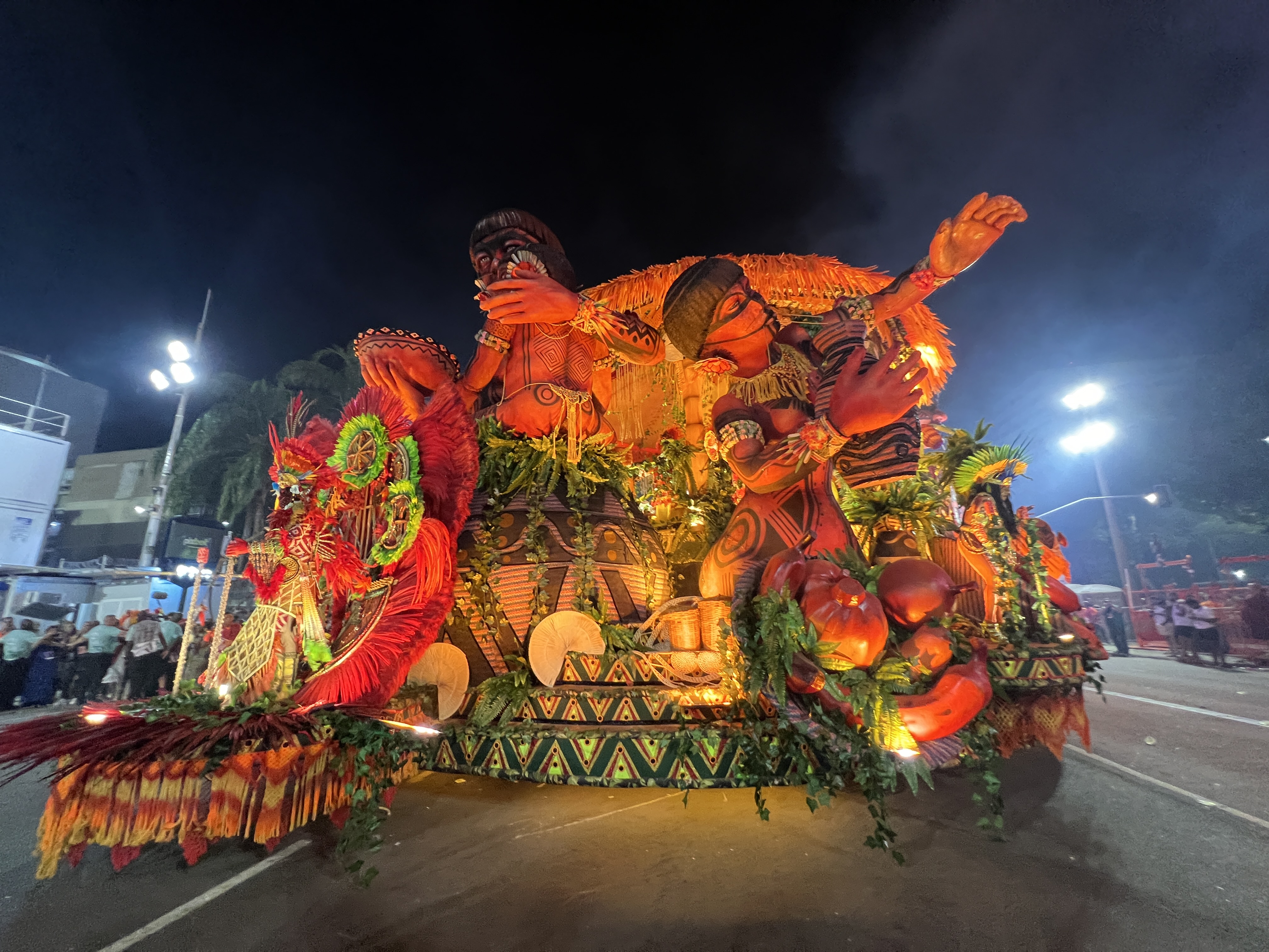 A parade float, depicting Yanomami people atop cultural symbols like woven baskets, moves down a paved road to the Sambodrome in Rio de Janeiro.