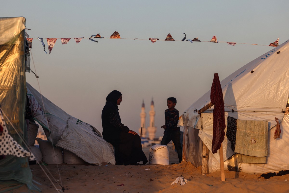 A boy walks past a Palestinian woman sitting at a camp for displaced people backdropped by the minarets of a mosque, on the first day of the Muslim holy fasting month of Ramadan, in Rafah