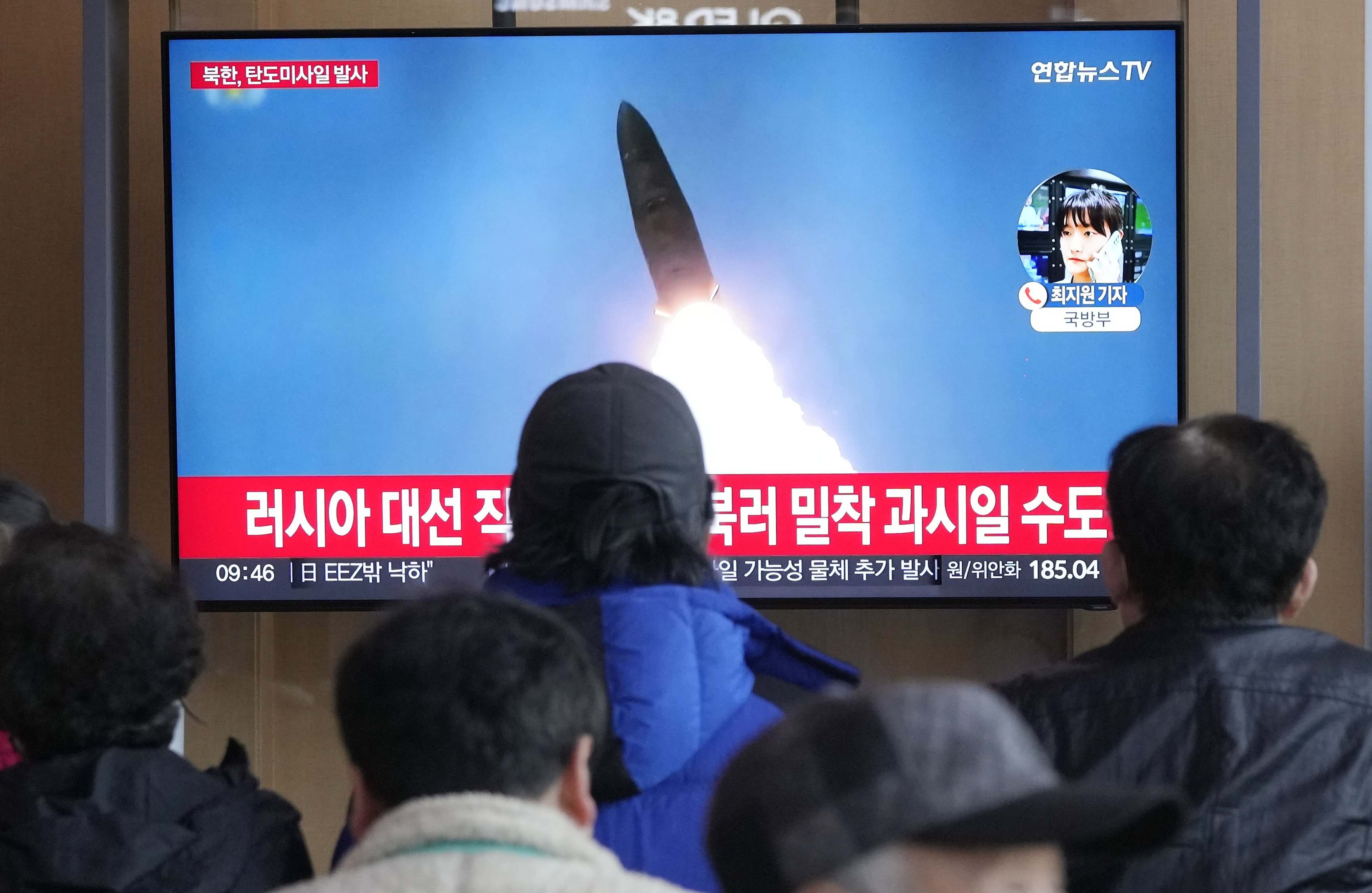 people watch a tv with a rocket launching and red news chyron