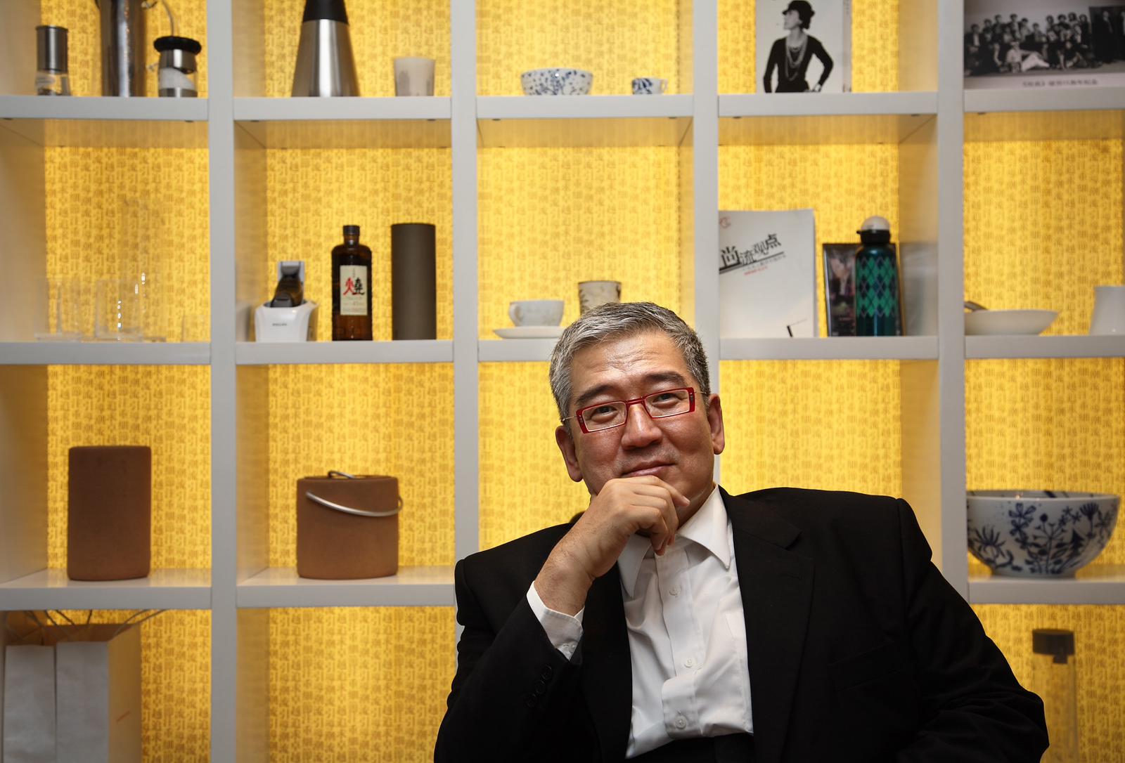 Rex How, He is seated in front of open shelving. He is smiling.