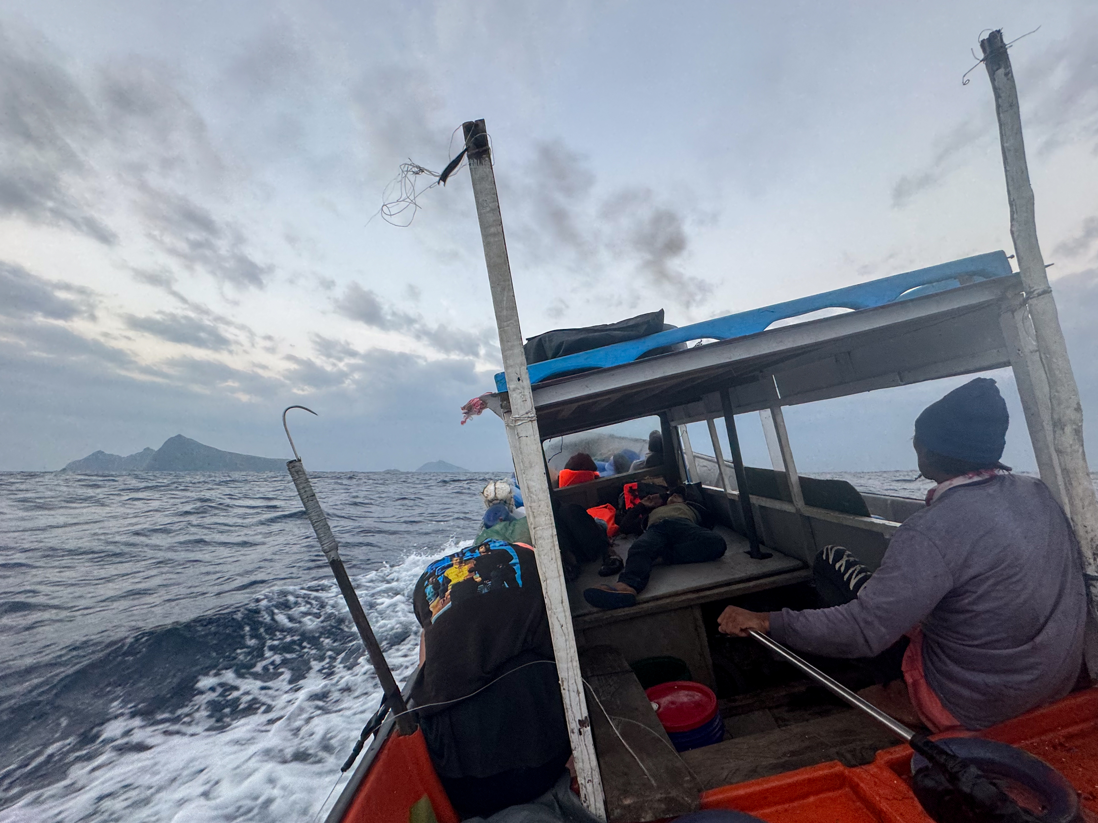 A fishing boat making its way to Mavulis. There are people in life vests towards the bow and a roof over some of the deck. The boat is listing to one side. A mountainous island can be seen ib the distance