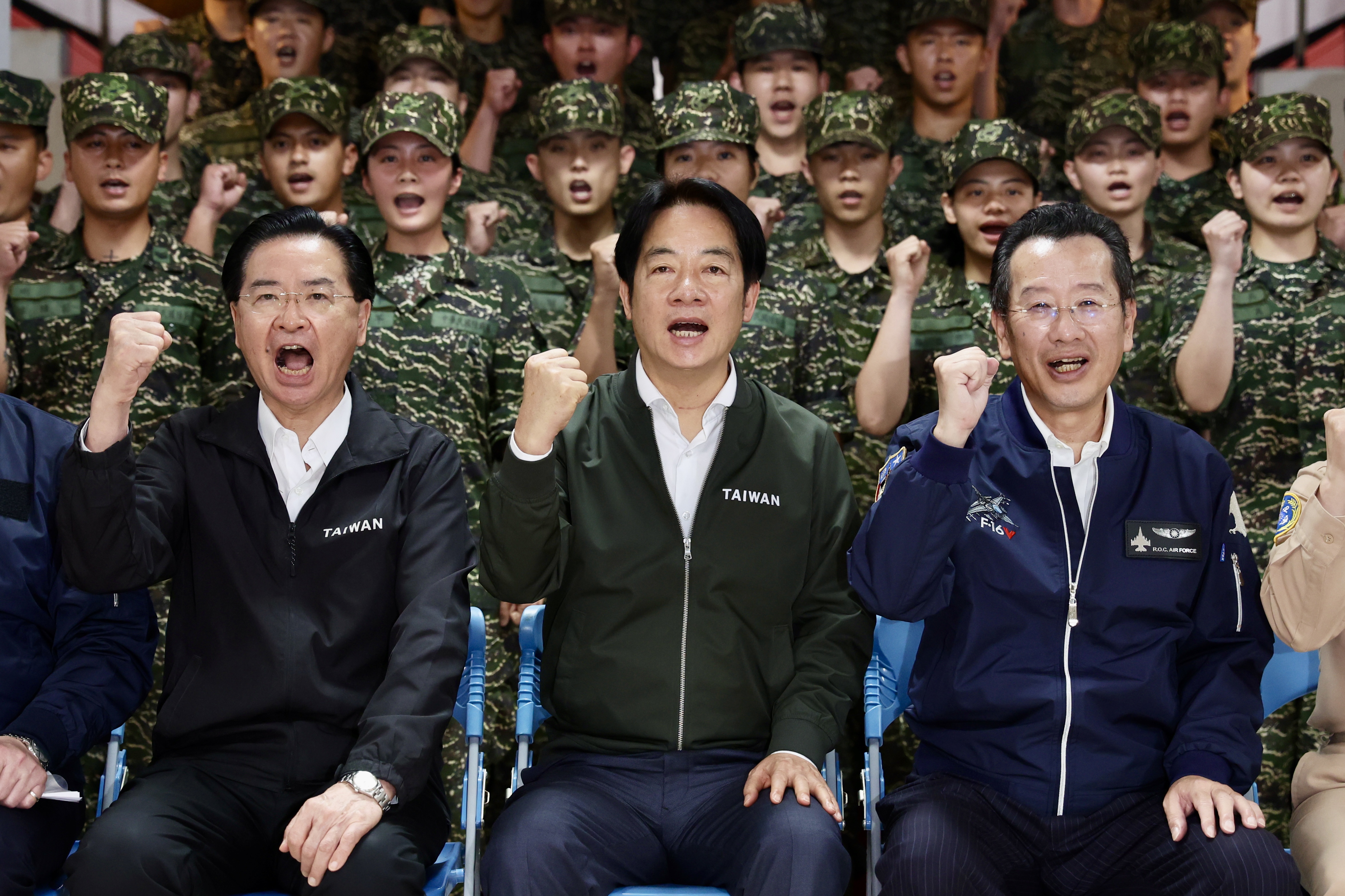 Taiwan's President William Lai Ching-te at a visit to a military base. He is seated with two officials and punching the air. Soldiers in uniform are behind them.