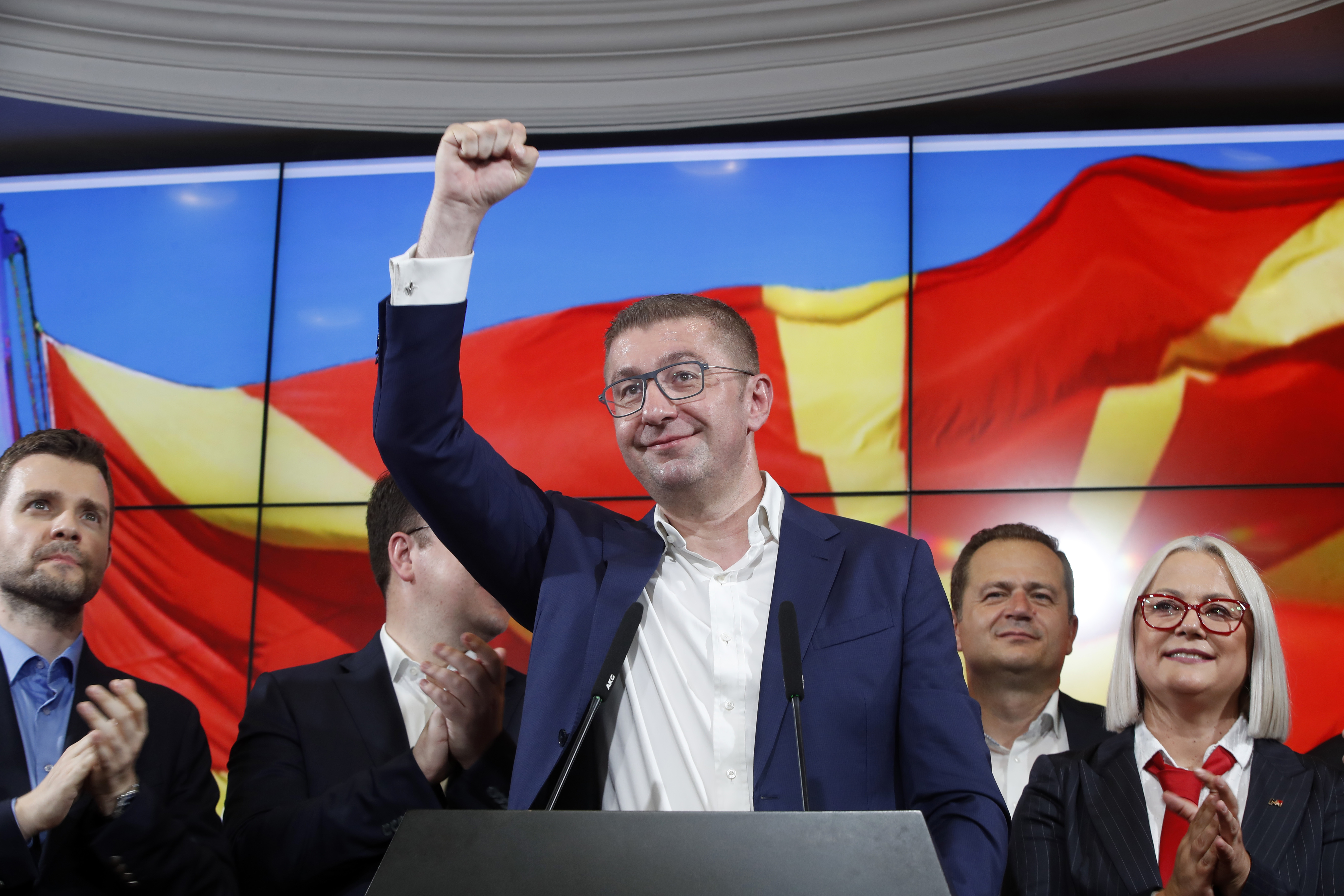 Hristijan Mickoski raises a fist in the air from behind a podium as he celebrates election-night results on a stage backed by screens.
