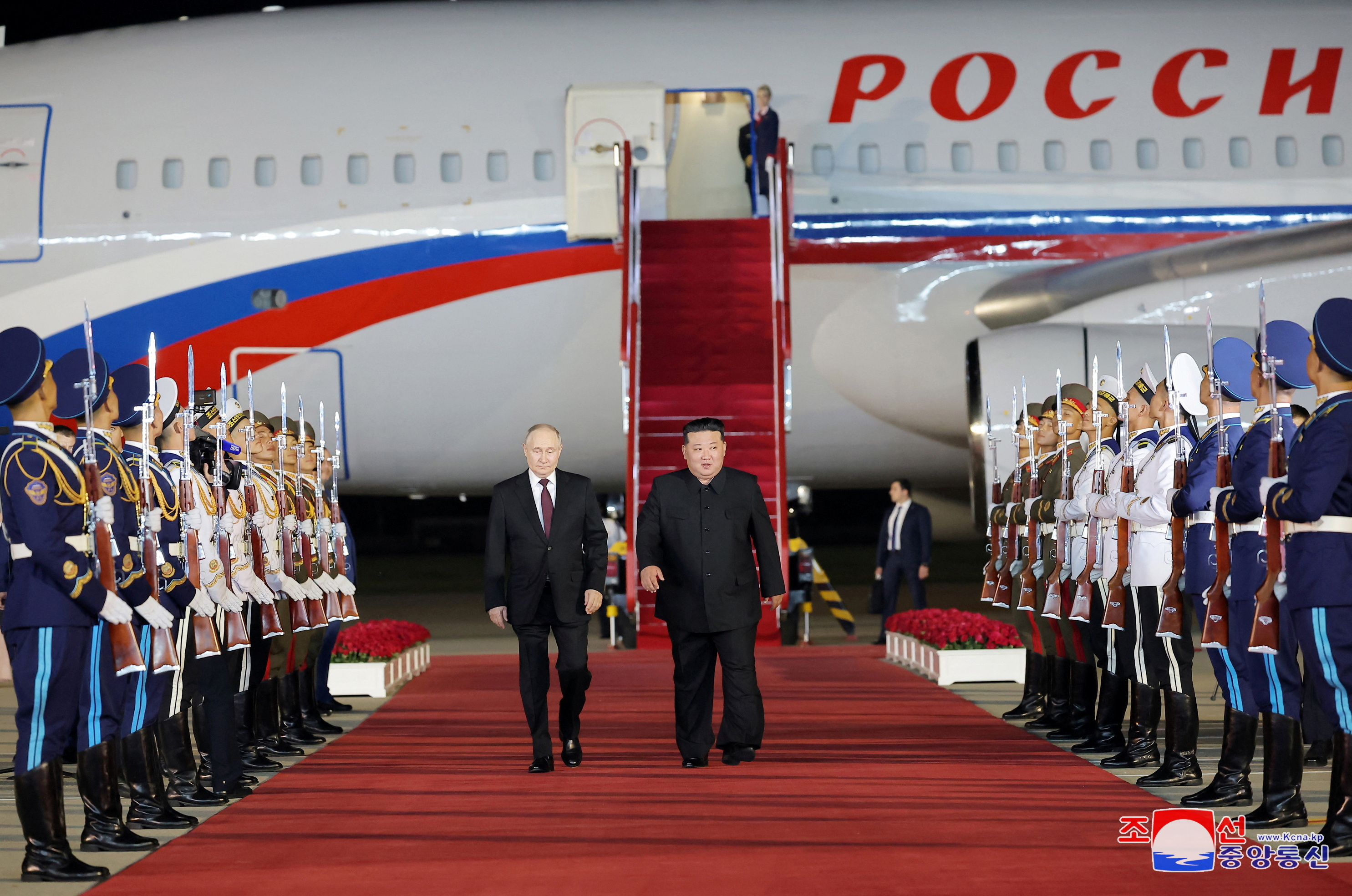 Vladimir Putin and Kim Jong Un walking down the red carpet at the airport. Putin's plane is behind him. Ceremonial guards are standing to attention on either side
