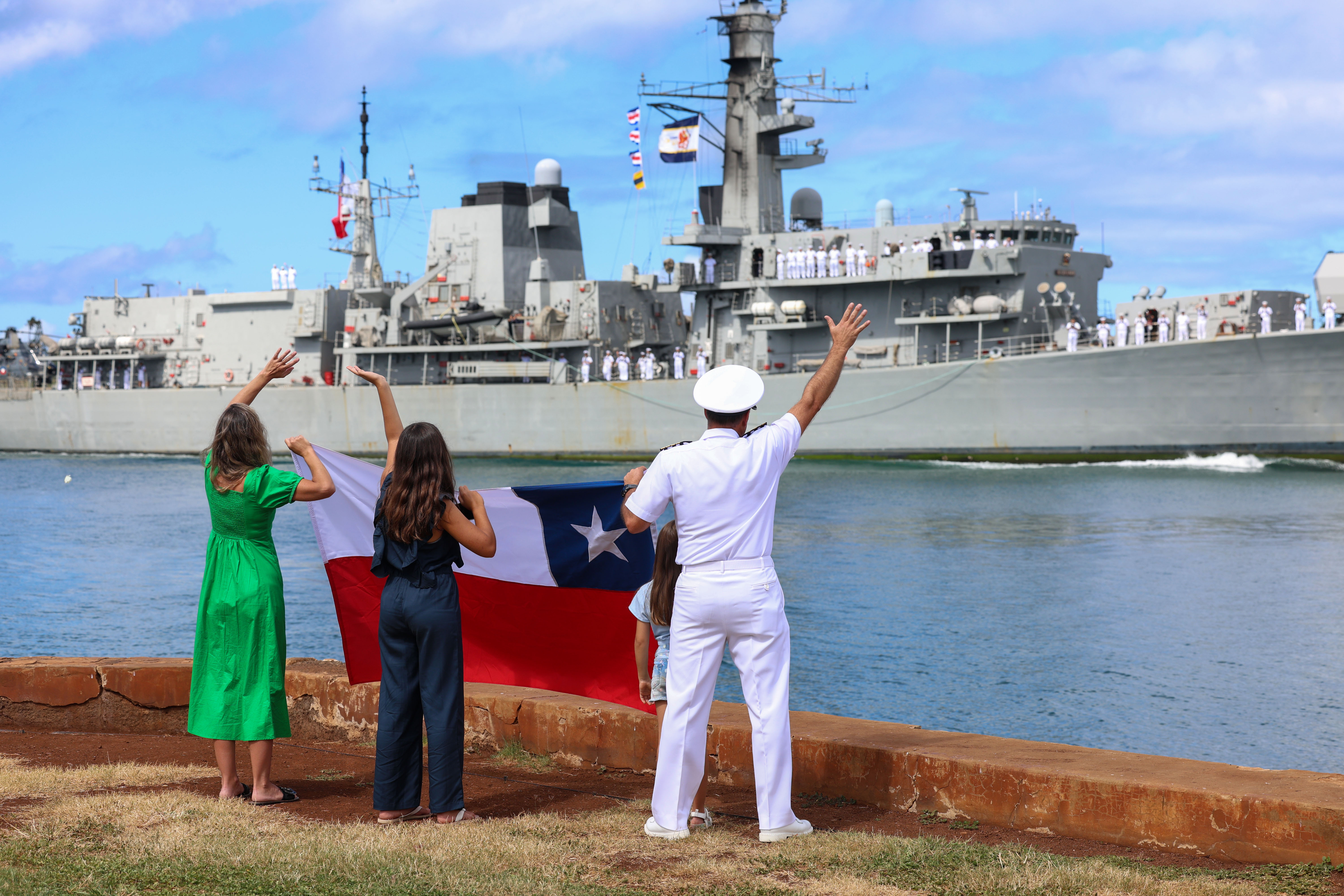 A Chilean navy captain and his family wave to a Chilean frigate as it arrives in Hawaii for the RIMPAC exercises. They are standing on the quay and holding a Chilean flag.