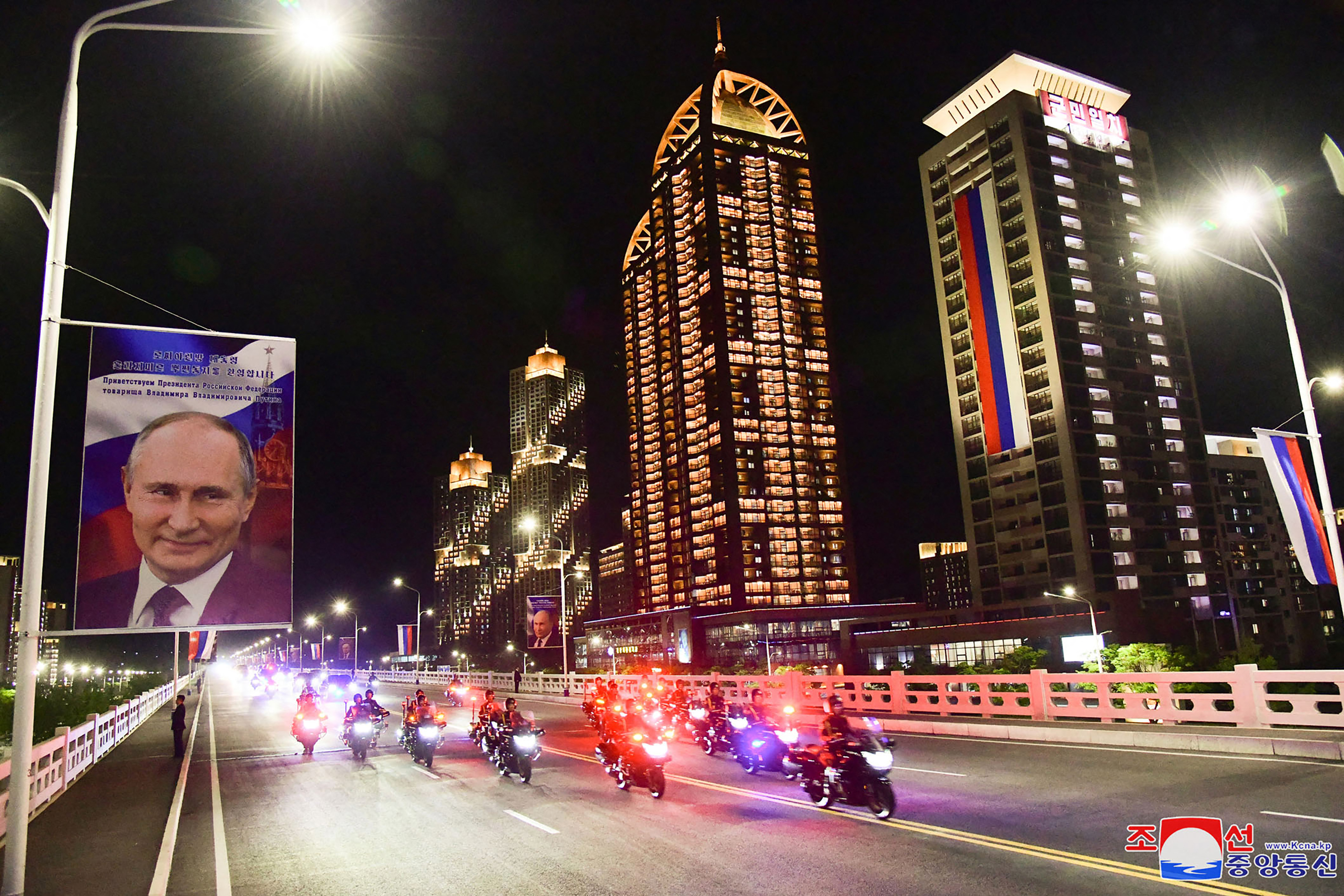 The motorcade travelling along a Pyongyang road. There are high rises behind with the lights on. Portraits of Putin have been put up along the side of the street.