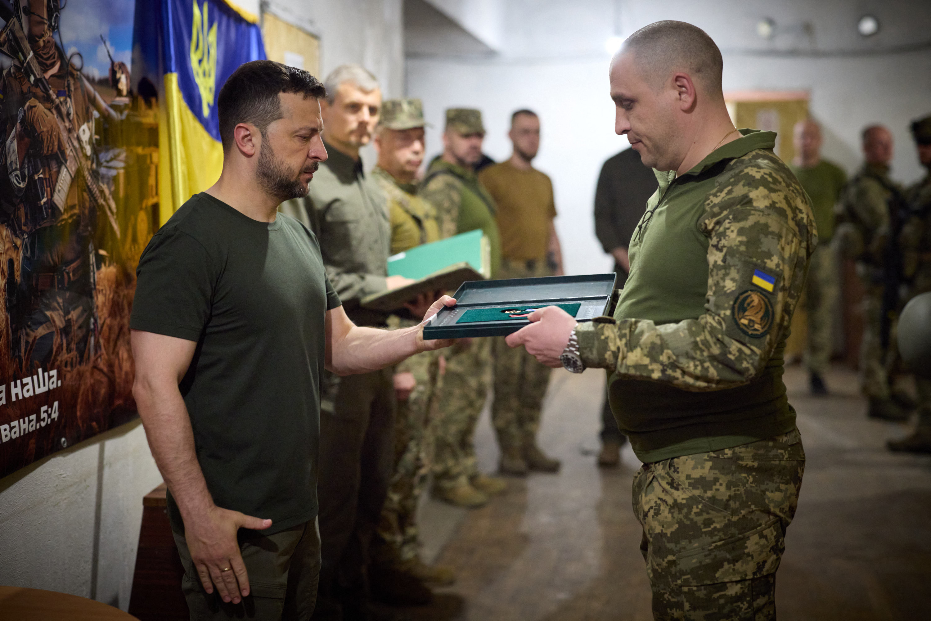 Volodymyr Zelenskyy giving an award to a Ukrainian soldier. He is wearing a dark green T-shirt and trousers. The soldier is in combat fatigues. Other soldiers are lined up to the side of Zelenskyy. There is a blue and yellow Ukrainian flag on the wall