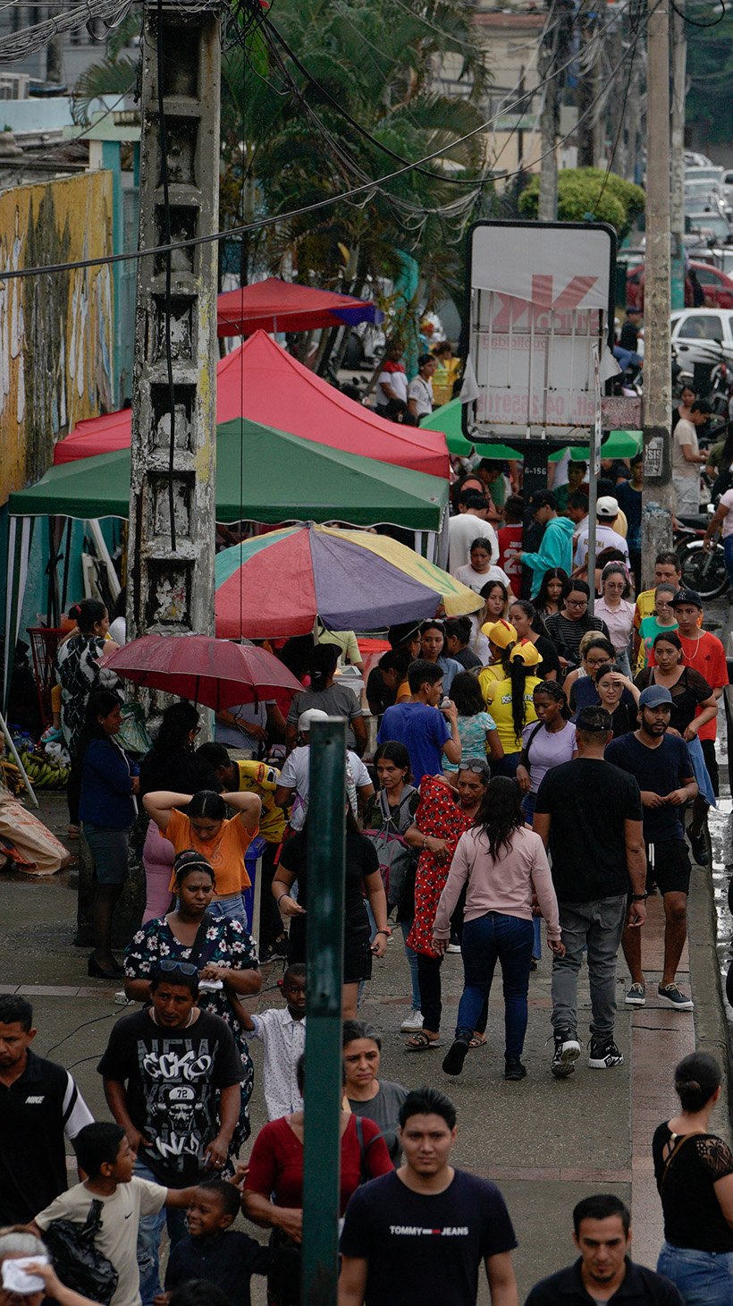 A street scene in Guayaquil, seen from above: cars and pedestrians navigating a busy street, moving past vendors with umbrellas.