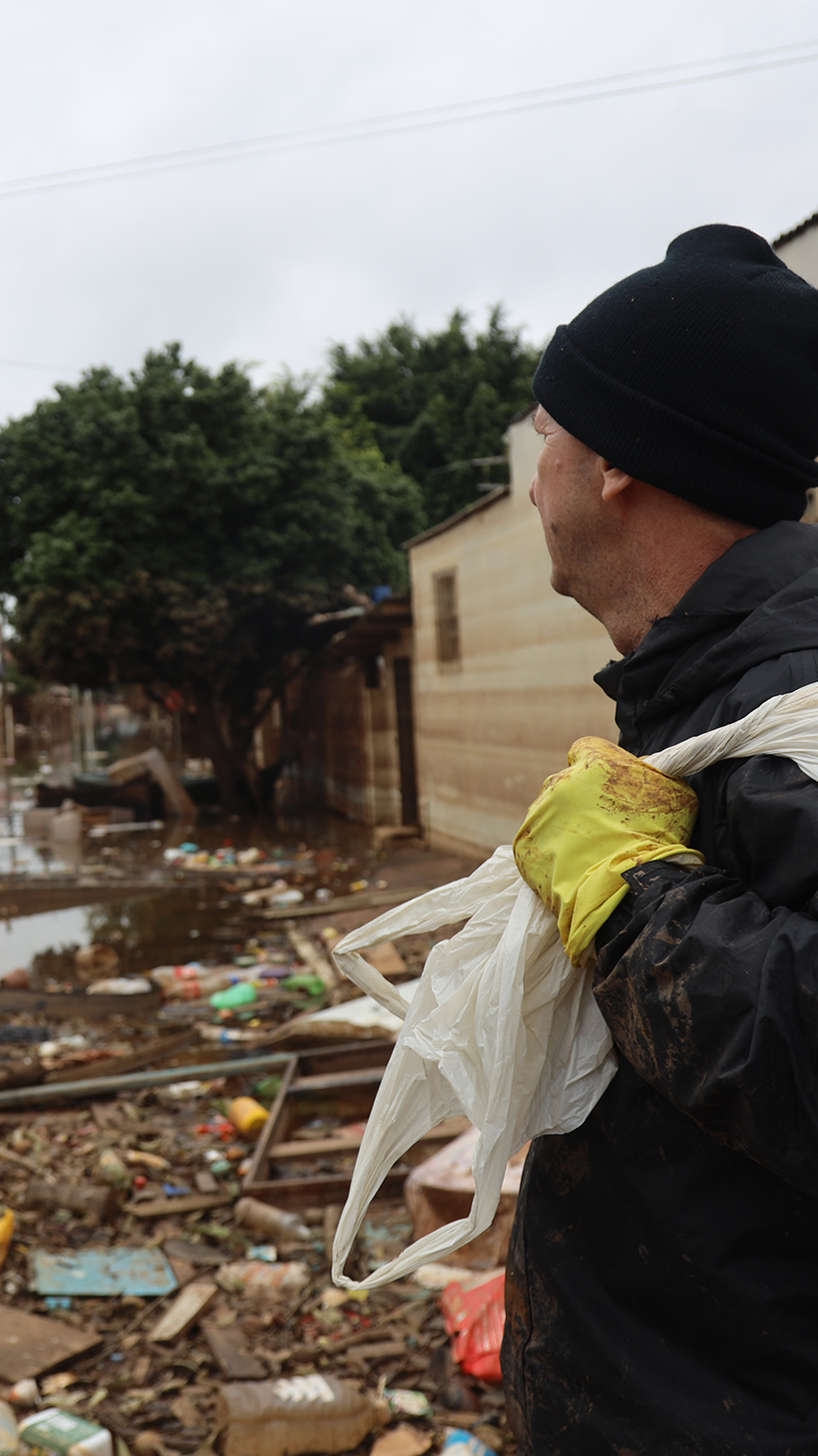 A man with a trash bag slung over his shoulder looks at a neighborhood devastated by floods, the ground still muddy and loose boards and detritus strewn through the streets.