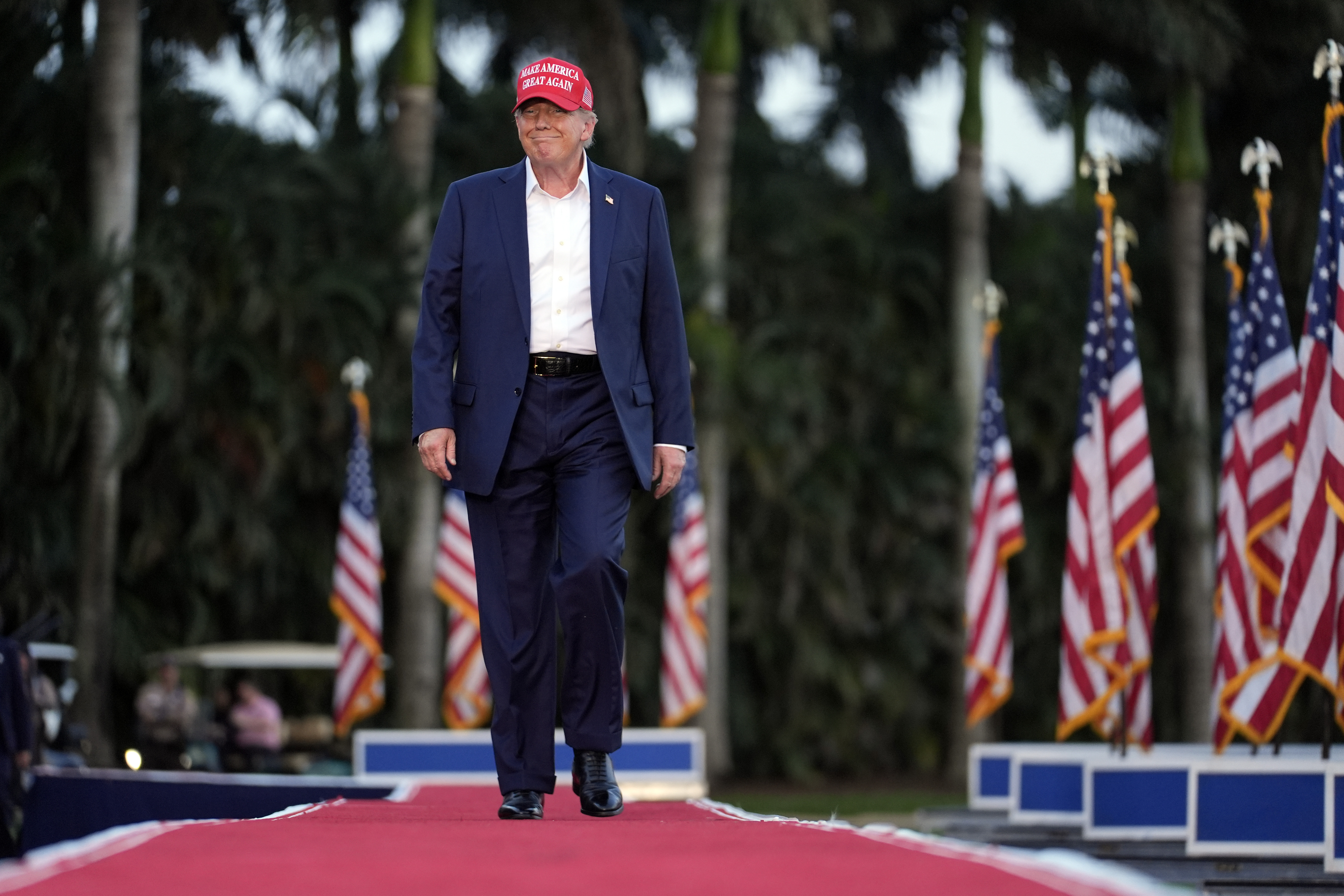 Donald Trump walks on a red carpet outdoors, past a row of US flags.