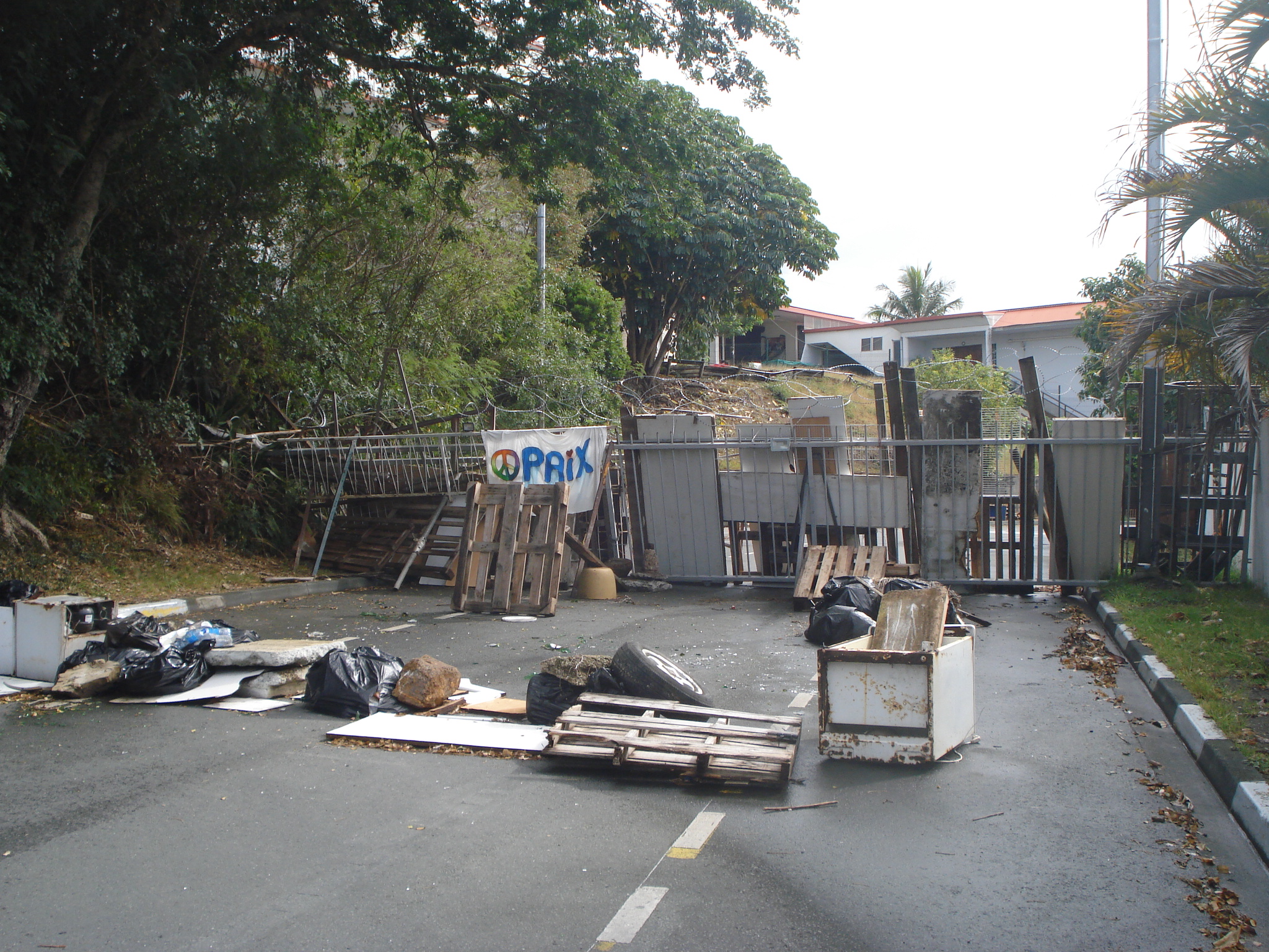 A barricade to a housing area in Noumea. The main barrier is made of railings, timber slats and wooden pallets. A sheet with the word 'Paix' (peace) has been hung from it. There are old fridges and other items lying on the road in front.