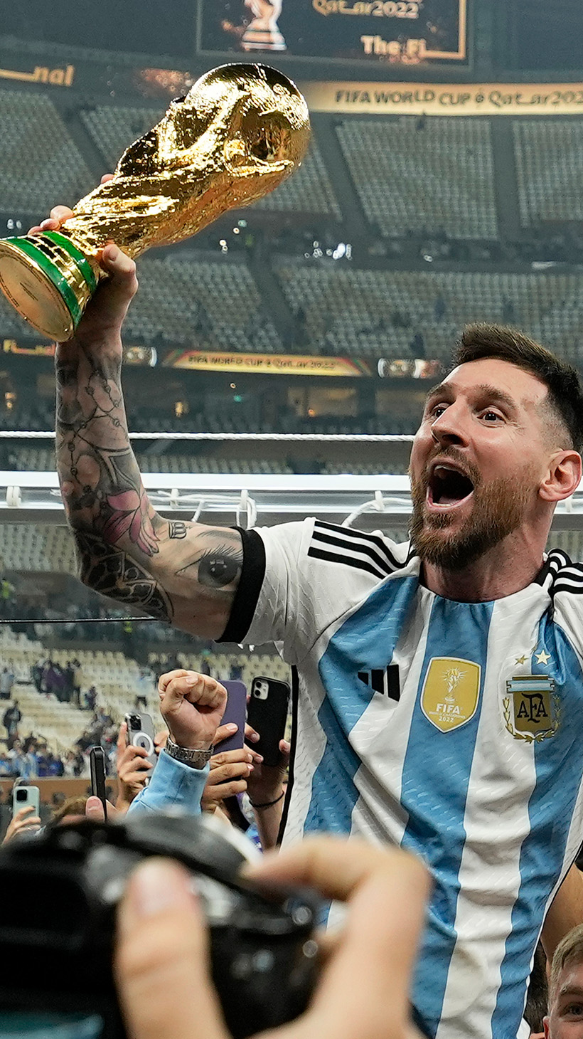 Lionel Messi, dressed in his blue and white Argentine stripes, is raised aloft by a crowd of supporters. In one hand, he carries the FIFA trophy.