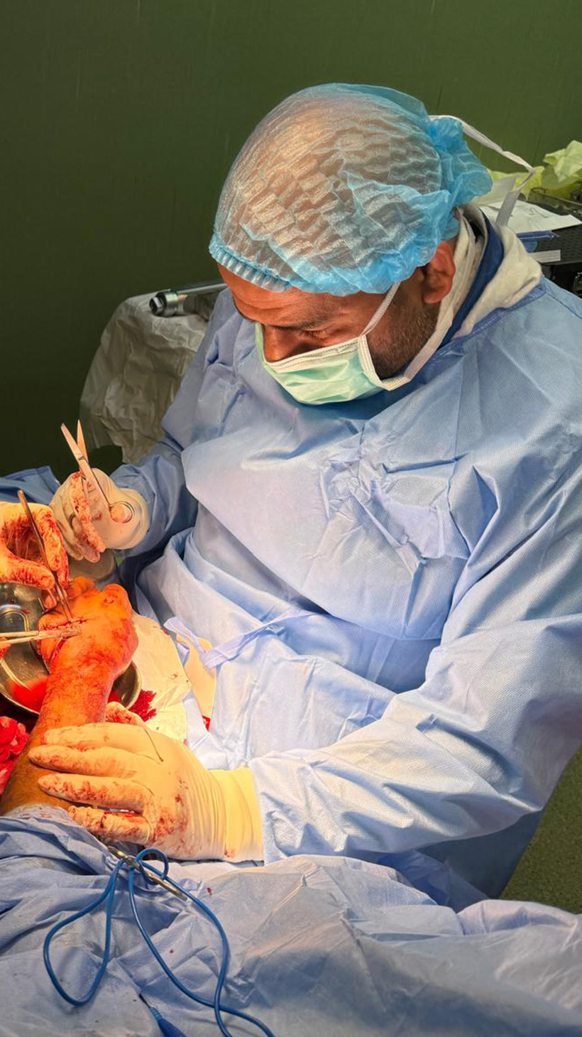 Omar Sabha and Jawad Khan, dressed in blue medical scrubs and hair nets, operate on a patient's hand, stitching it up. Blood is visible on the medical worker's gloves.