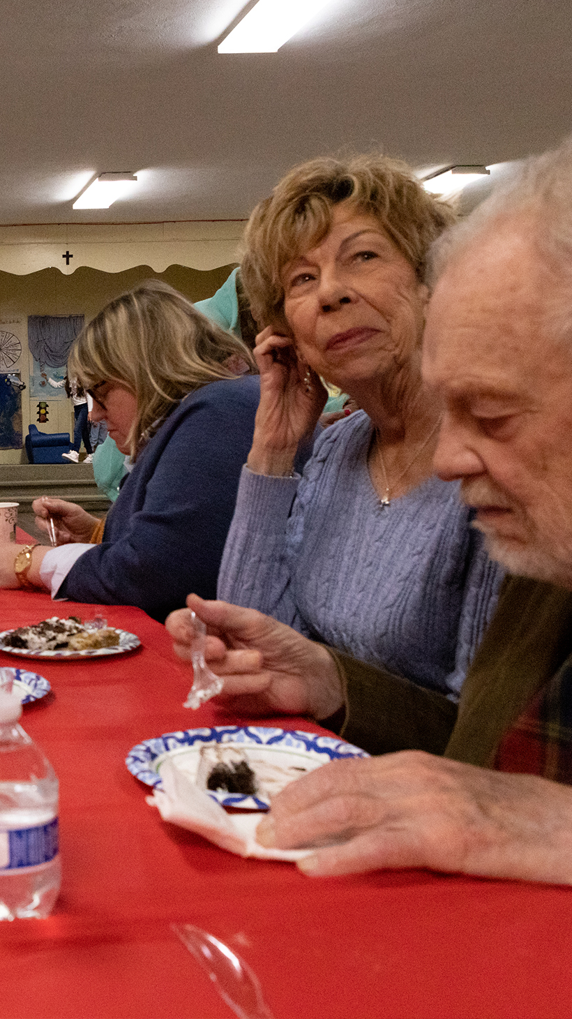 Parishioners gather around a long table with a red table cloth to eat cake off of paper plates.