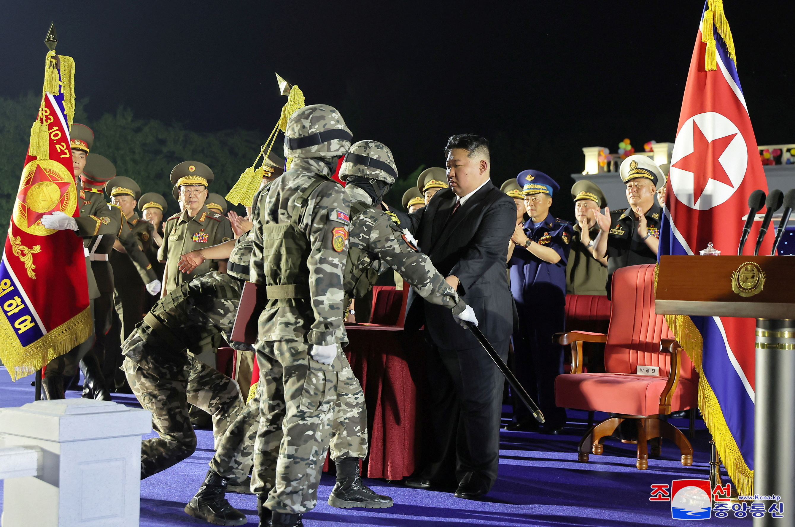 Kim Jong Un handing a flag to a soldier in combat uniform. They are on stage at a ceremony, Kim is dressed in black. The soldiers are in combat uniform. There are other officials nearby, 