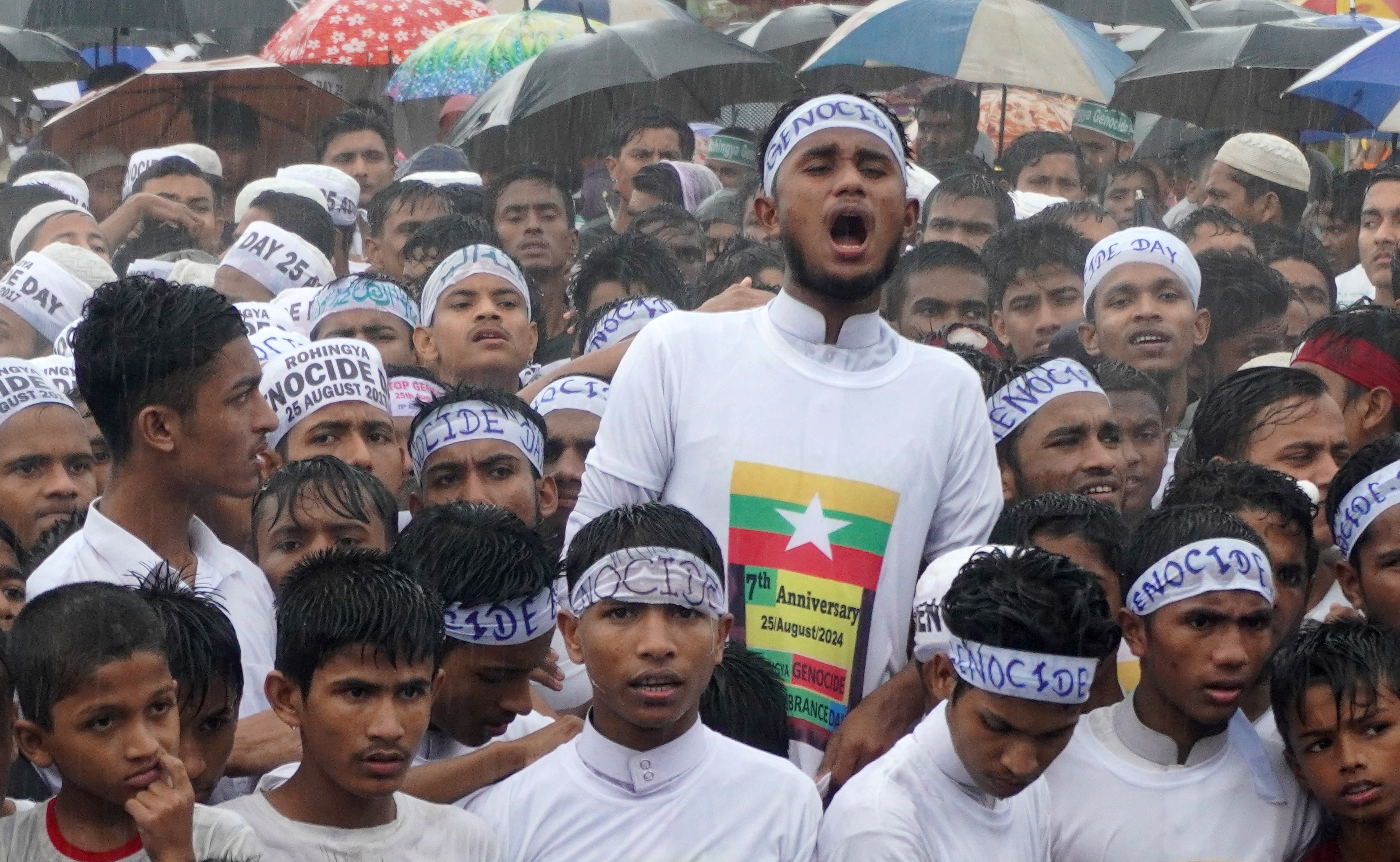 Young Rohingya men at a protest. They are dressed in white and have bandanas tied around their heads reading 'Genocide' One is wearing a T-shirt with the Myanmar flag. It is raining.