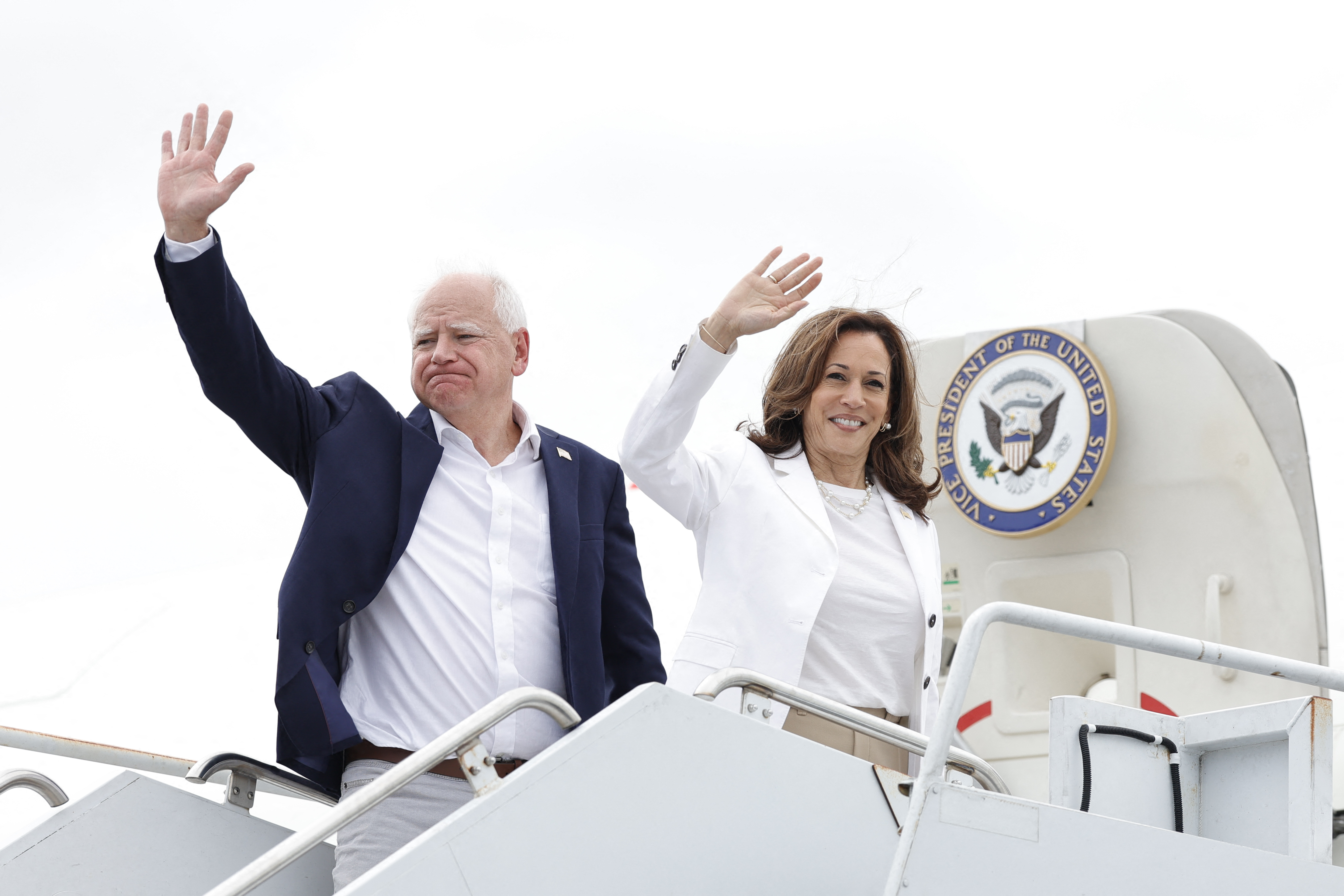Kamala Harris and Tim Walz about to board Air Force Two. They are standing at the top of the steps and waving.
