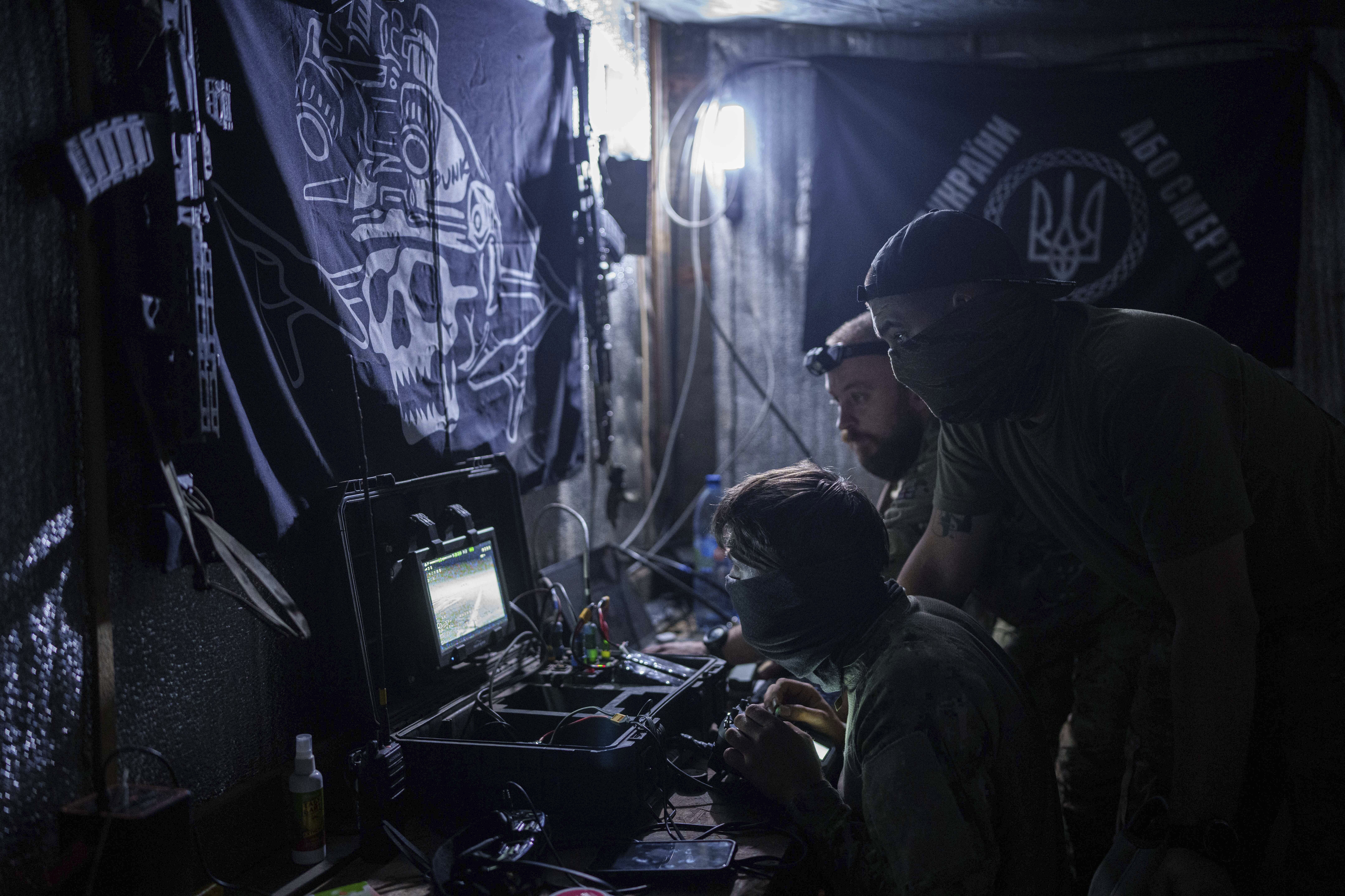 Ukrainian soldiers around a desk controlling a drone. The place is very dark. There's a black banner on the wall in front of them.
