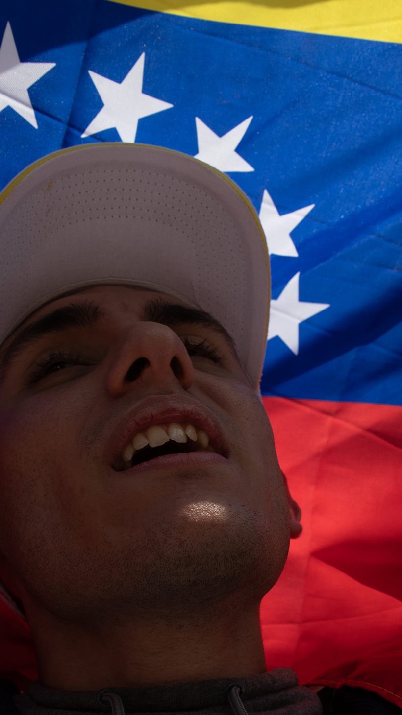 A man in a baseball cap raises a Venezuelan flag above his head, its breadth blocking out the sun behind him.