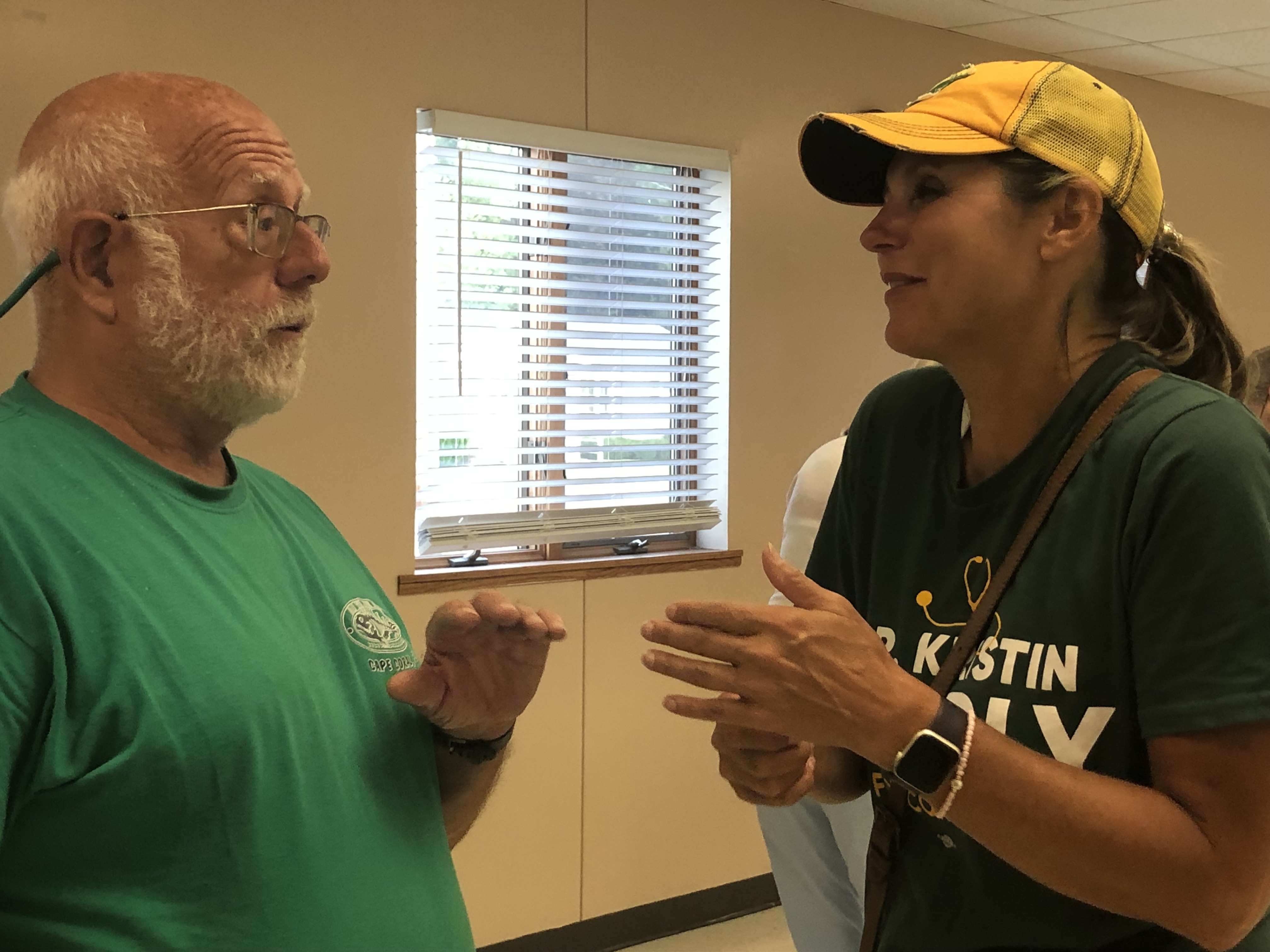 A woman in a yellow ball cap speaks animatedly to a man in a yellow room.