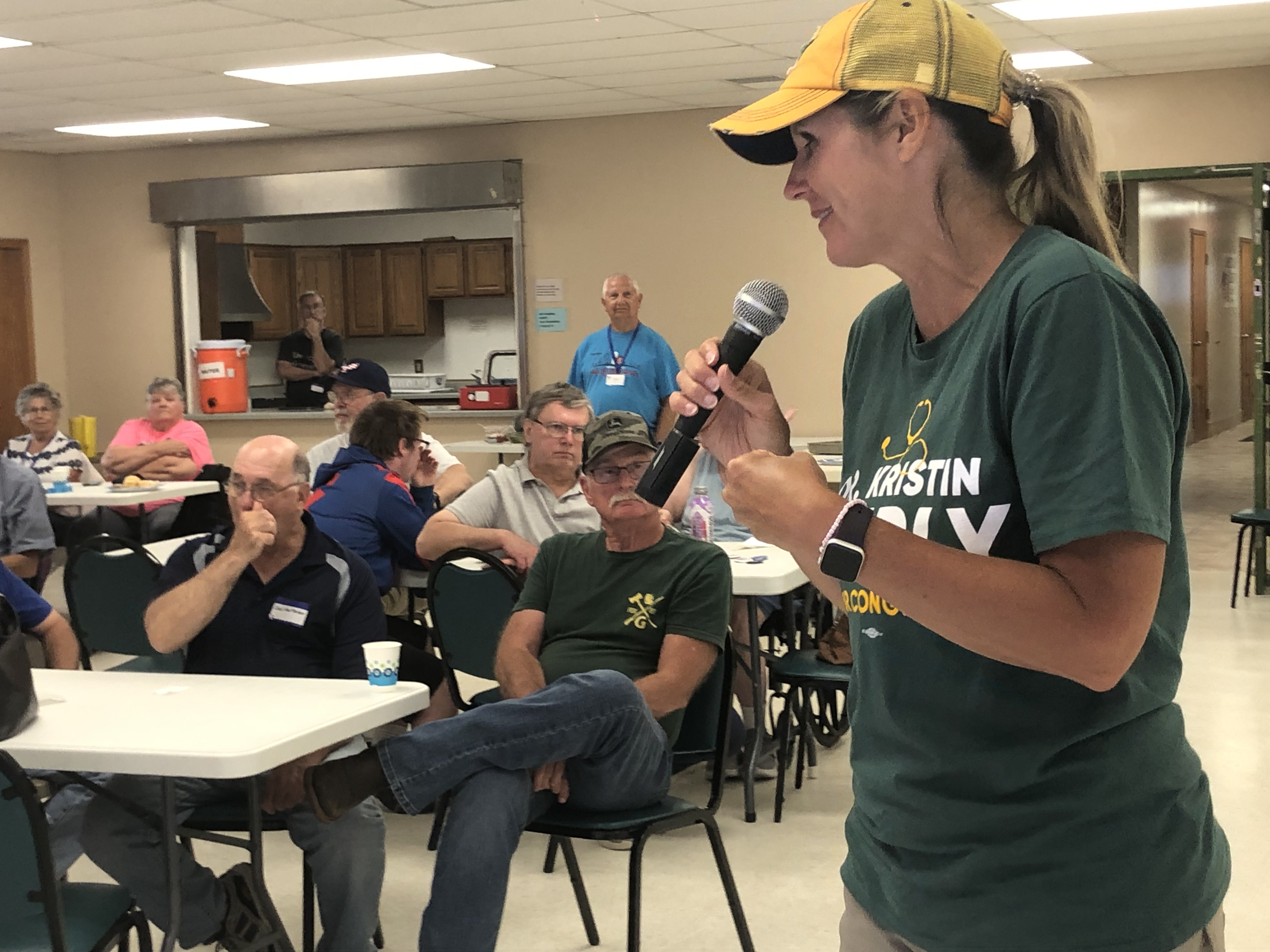 Kristin Lyerly, dressed in a yellow baseball cap and green shirt, speaks into a microphone in front of a room of voters.