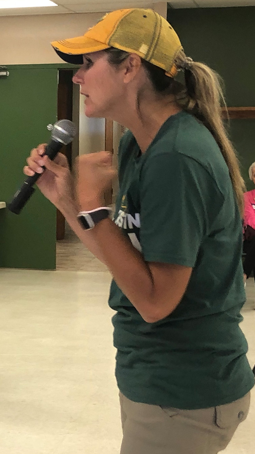 Kristin Lyerly, dressed in a yellow baseball cap and green shirt, speaks into a microphone in front of a room of voters.