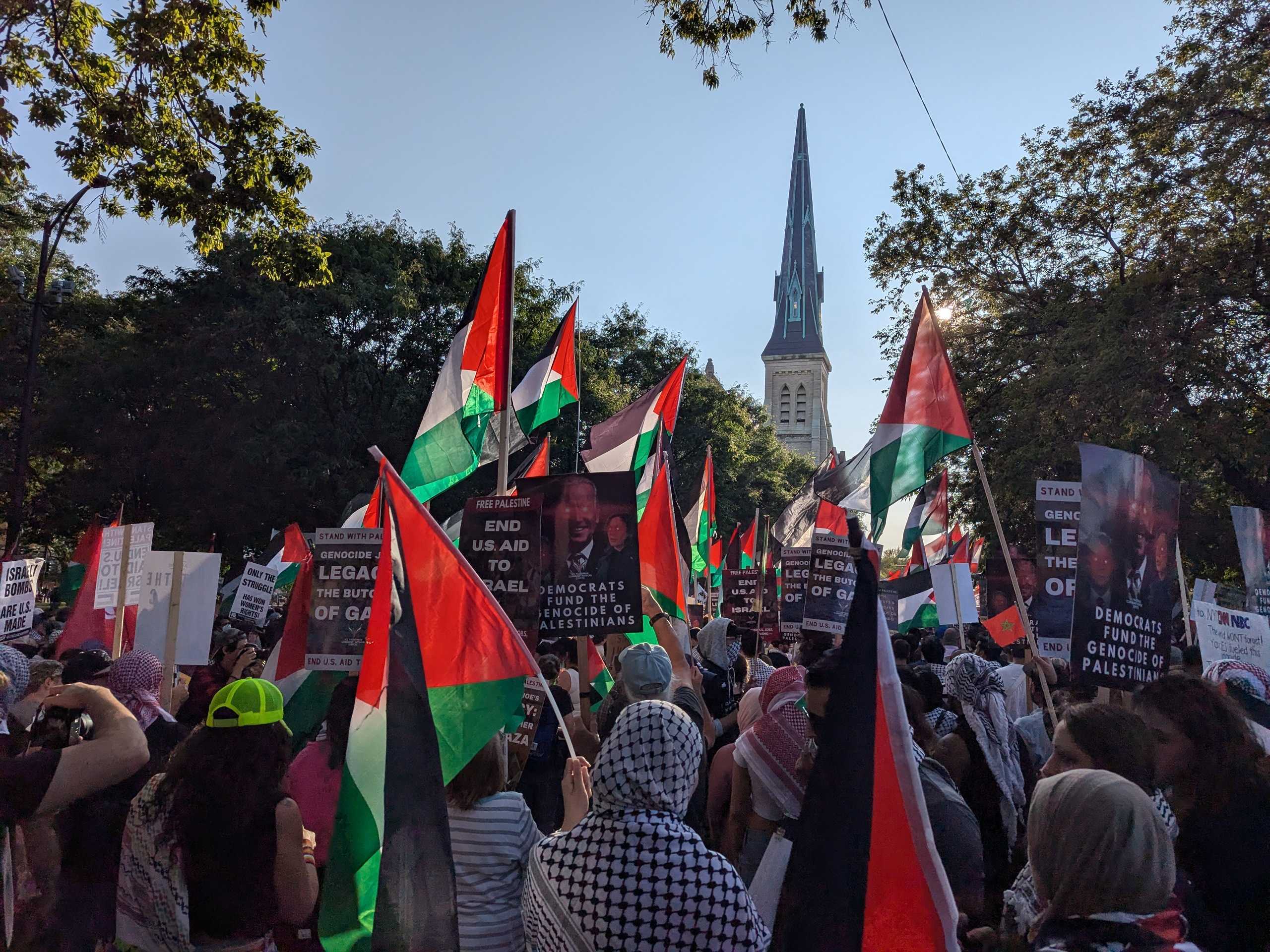 Protesters march down a Chicago street waving Palestinian flags. In the distance, a church spire rises above the treeline.