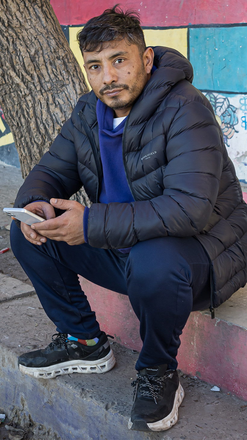 Waldemar Cubilla sits on a street curb, dressed in a quilted, hooded jacket. In his hands is a cellphone.