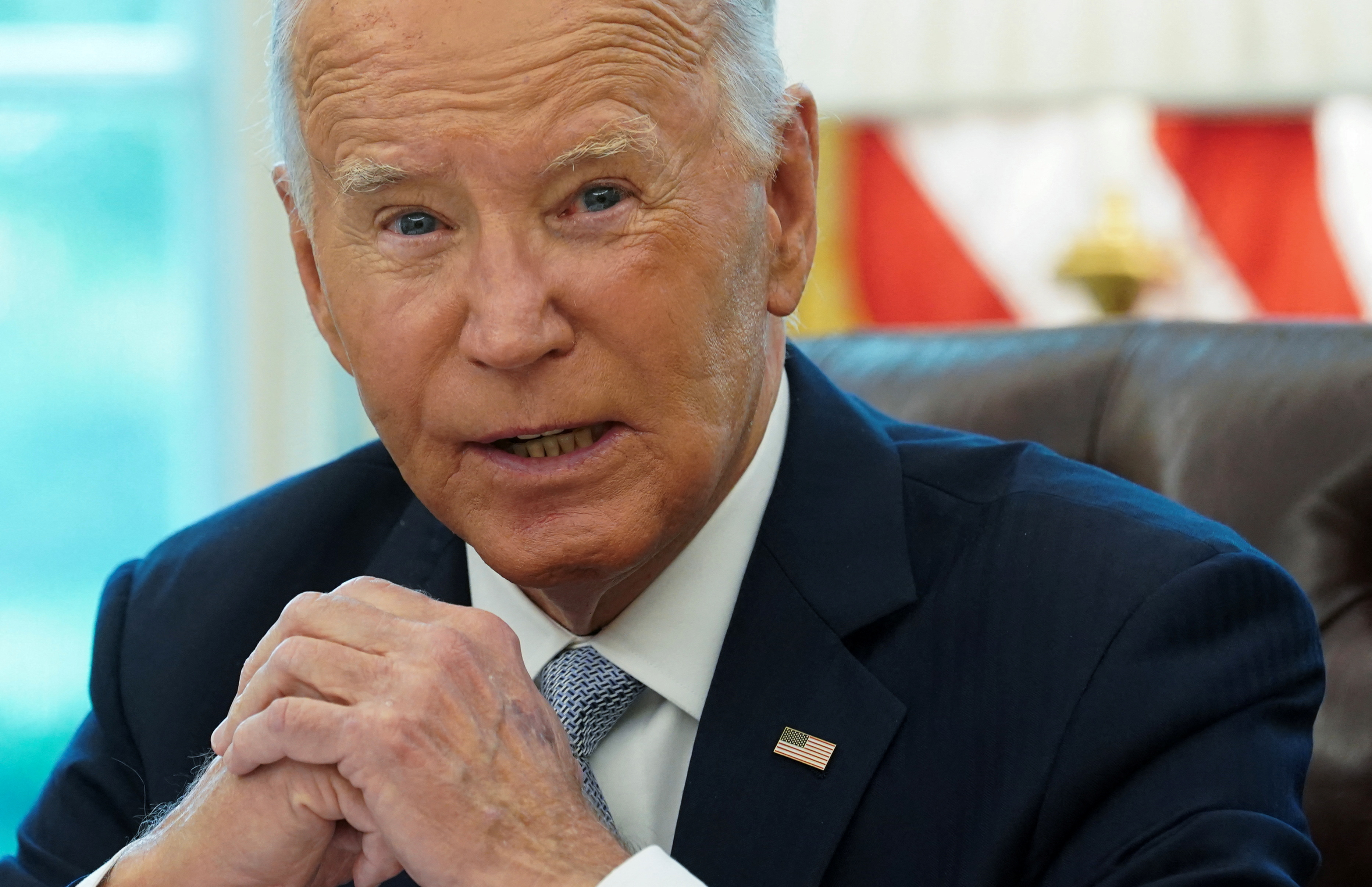 A head and shoulders photo of Joe Biden. He is sitting at his desk and has his handsl clasped in front of him