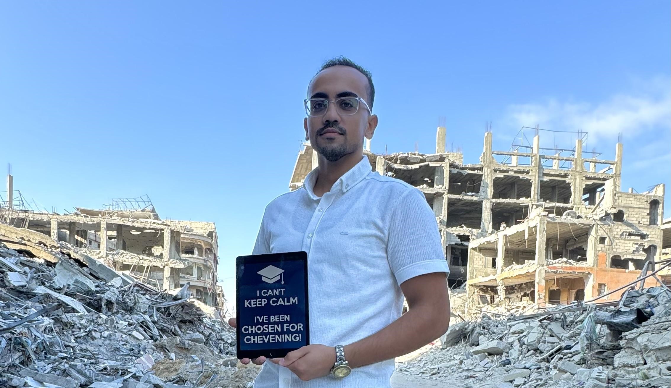 a photo of a man holding a sign saying I can't keep calm I was awarded Chevening is seen standing and smiling in front of rubble