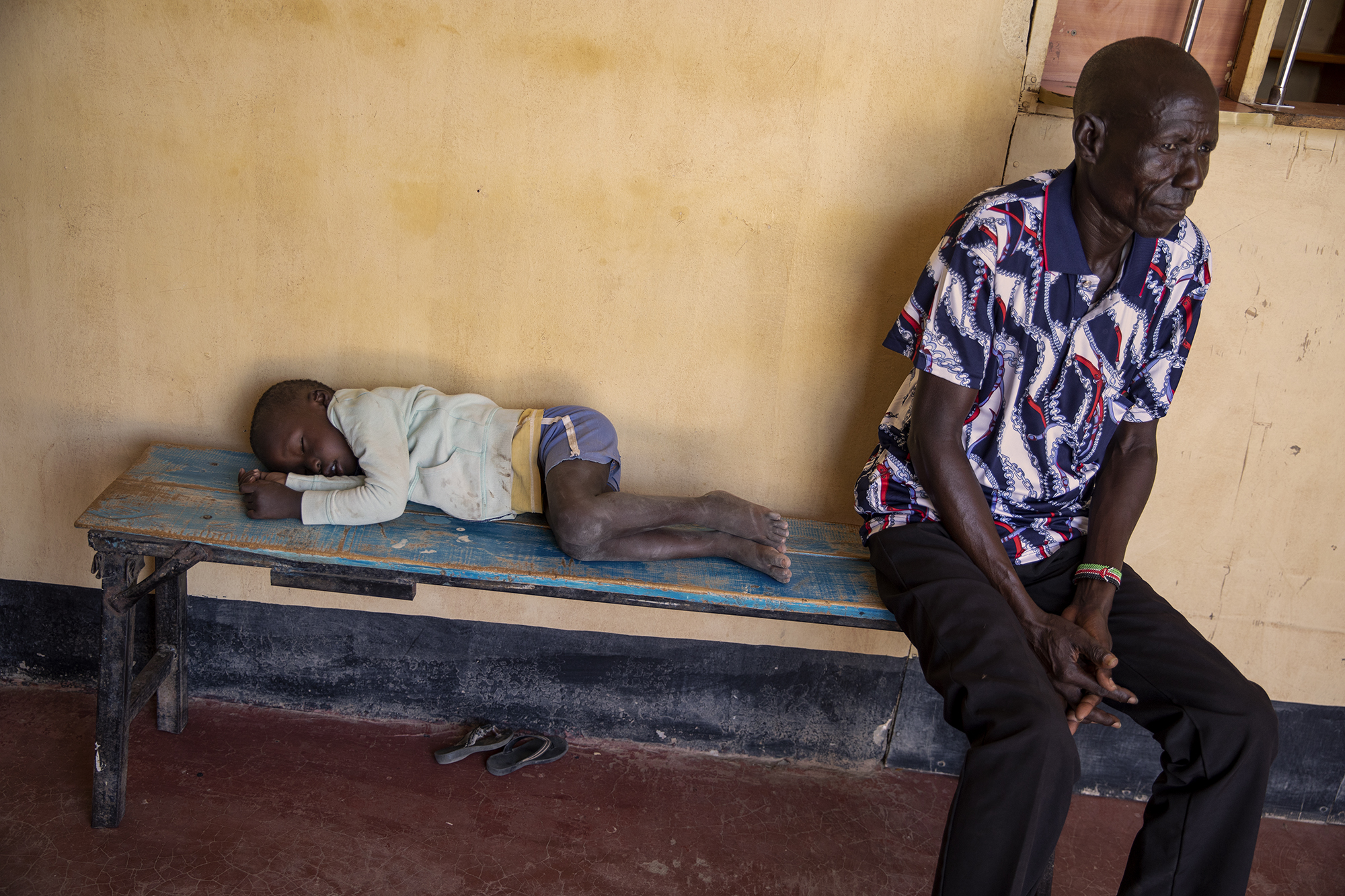 A child probably suffering from malaria sleeps on a bench, while a man waits his turn inside the Kalokol Gok Health Centre, located in Turkana County, northwest Kenya on October 16, 2024. Children often go fetching water with their mothers in areas where stagnant pools of water form and mosquitoes that carry the malaria parasite proliferate. Over the past three years Kenya has suffered from intense drought, partly caused by global warming, which also provides favourable conditions for the development of these mosquitoes. [Alessio Paduano/Al Jazeera]