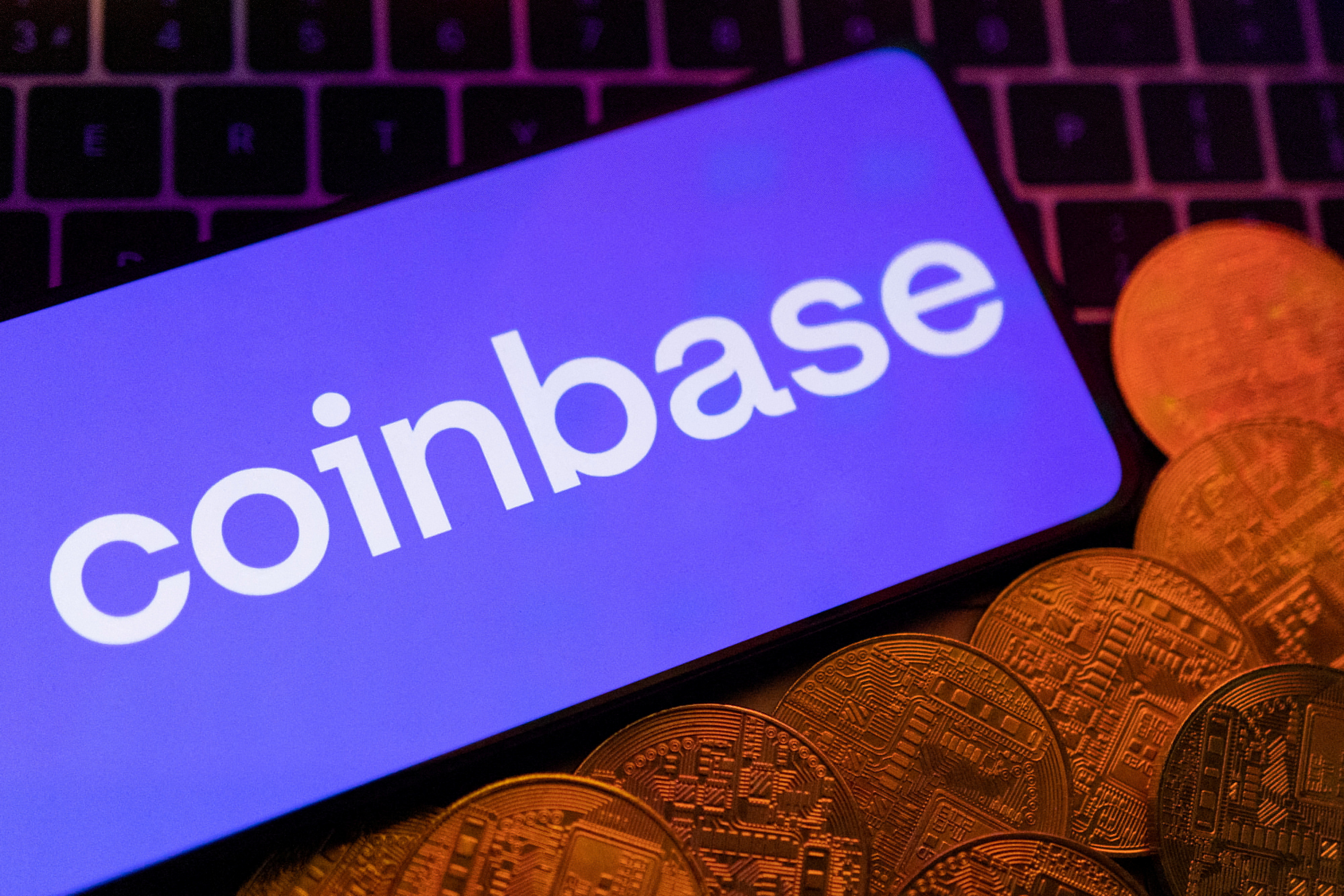 A smartphone showing the Coinbase logo rests on a keyboard beside metal coins stamped with cryptocurrency symbols.