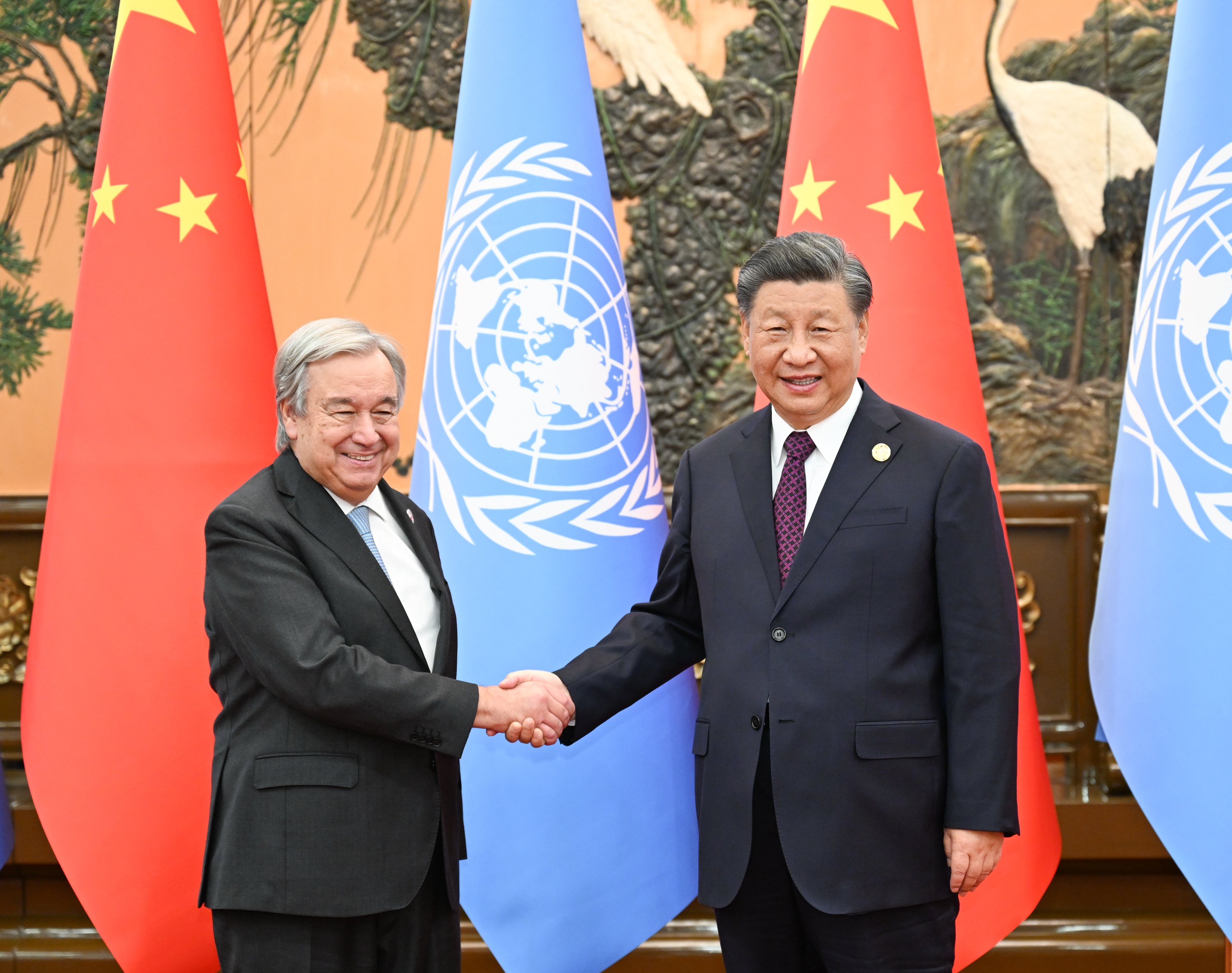 Chinese President Xi Jinping shakes hands with United Nations Secretary-General Antonio Guterres as they smile for the camera, with the Chinese and UN flags behind them.