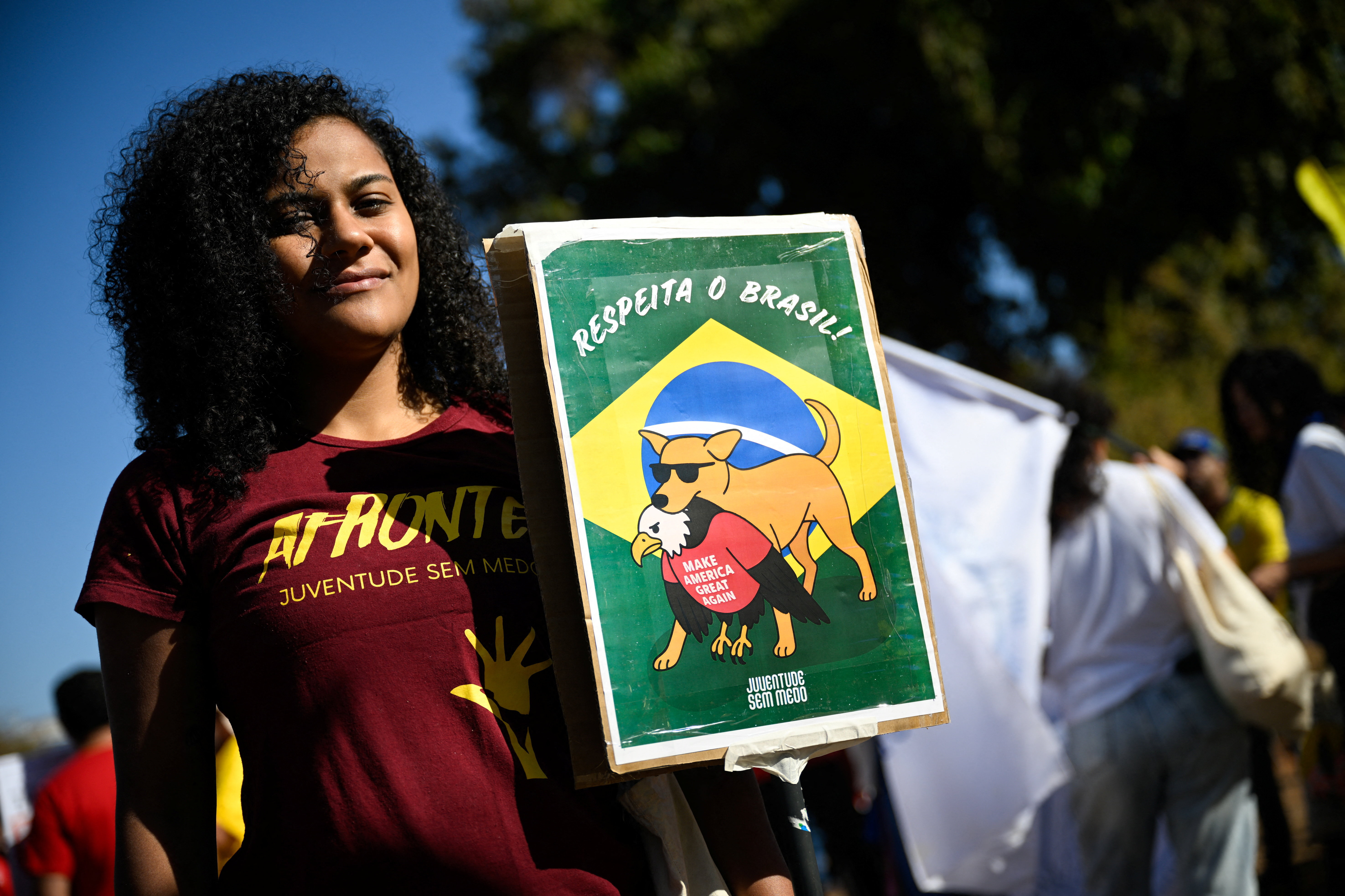 A protester holds up a sign that shows the Brazilian flag and a sunglasses-wearing dog biting an eagle with a red "Make America Great Again" shirt