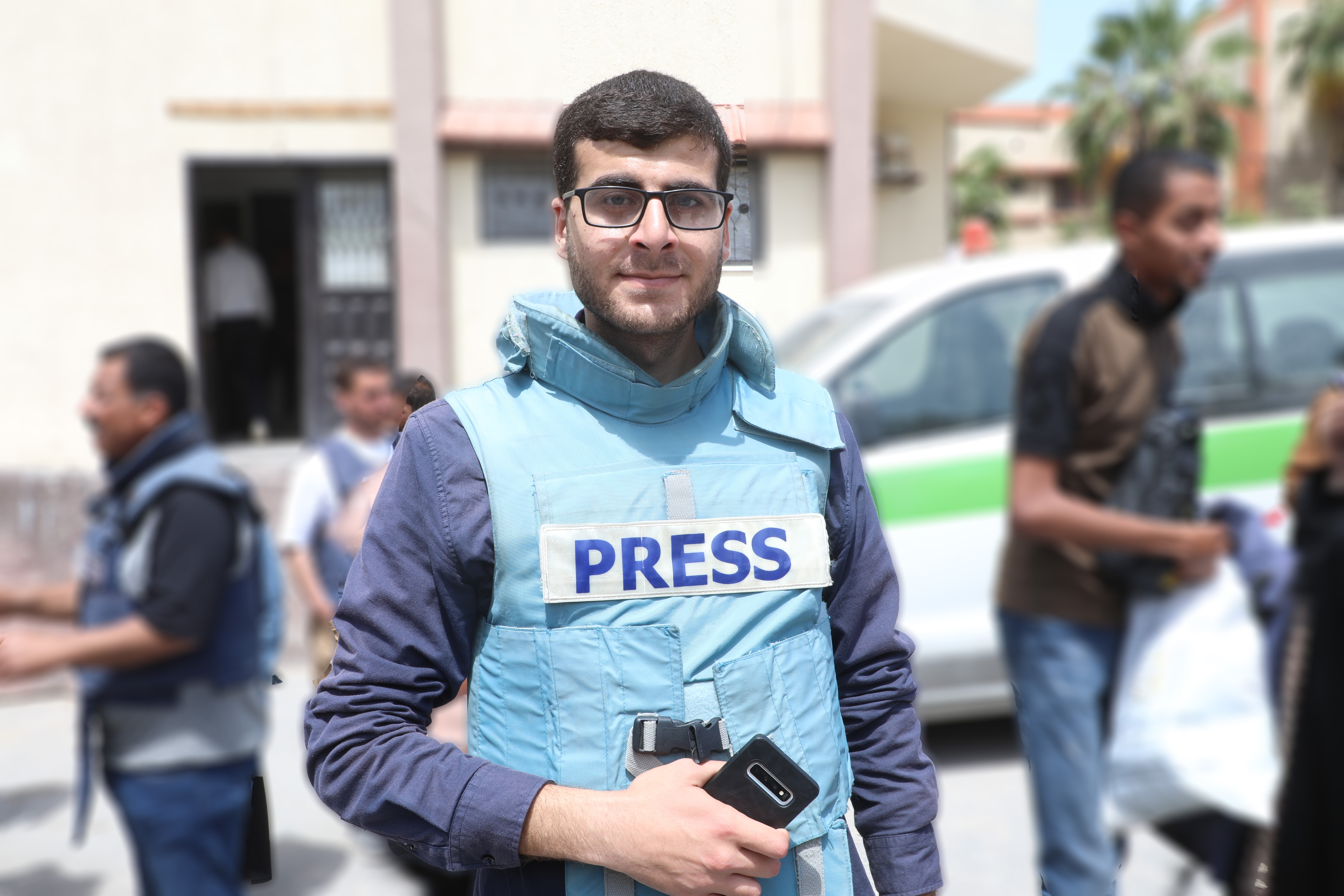 The author, dark-haried, wearing glasses, out reporting, wearing his press vest and holding his phone