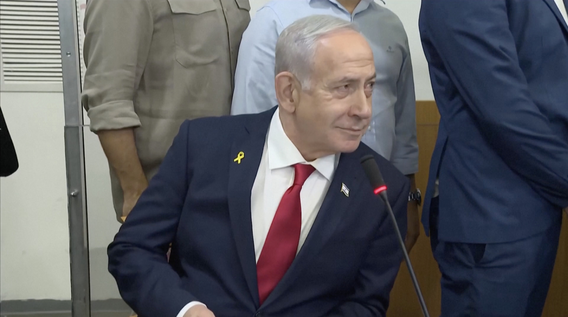 Israeli Prime Minister Benjamin Netanyahu seated, wearing a dark suit, white shirt, red tie, and yellow ribbon and Israeli flag pins on his lapel, with a microphone in front of him.