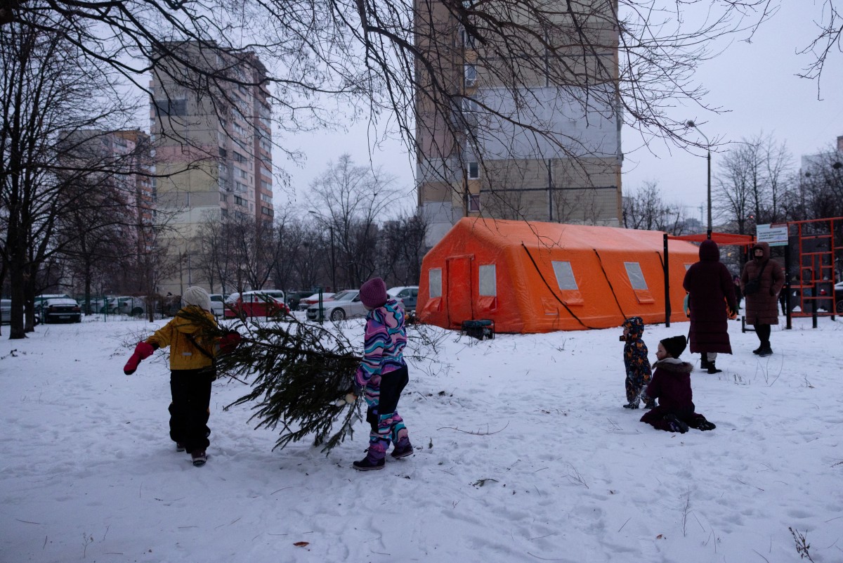 Children play with a Christmas tree by a tent of Point of Invincibility in Kyiv on January 11, 2026, where people can charge devices and warm themself, following Russian missile and drone attacks on Ukrainian energy infrastructure amid the Russian invasion in Ukraine. (Photo by Tetiana DZHAFAROVA / AFP)