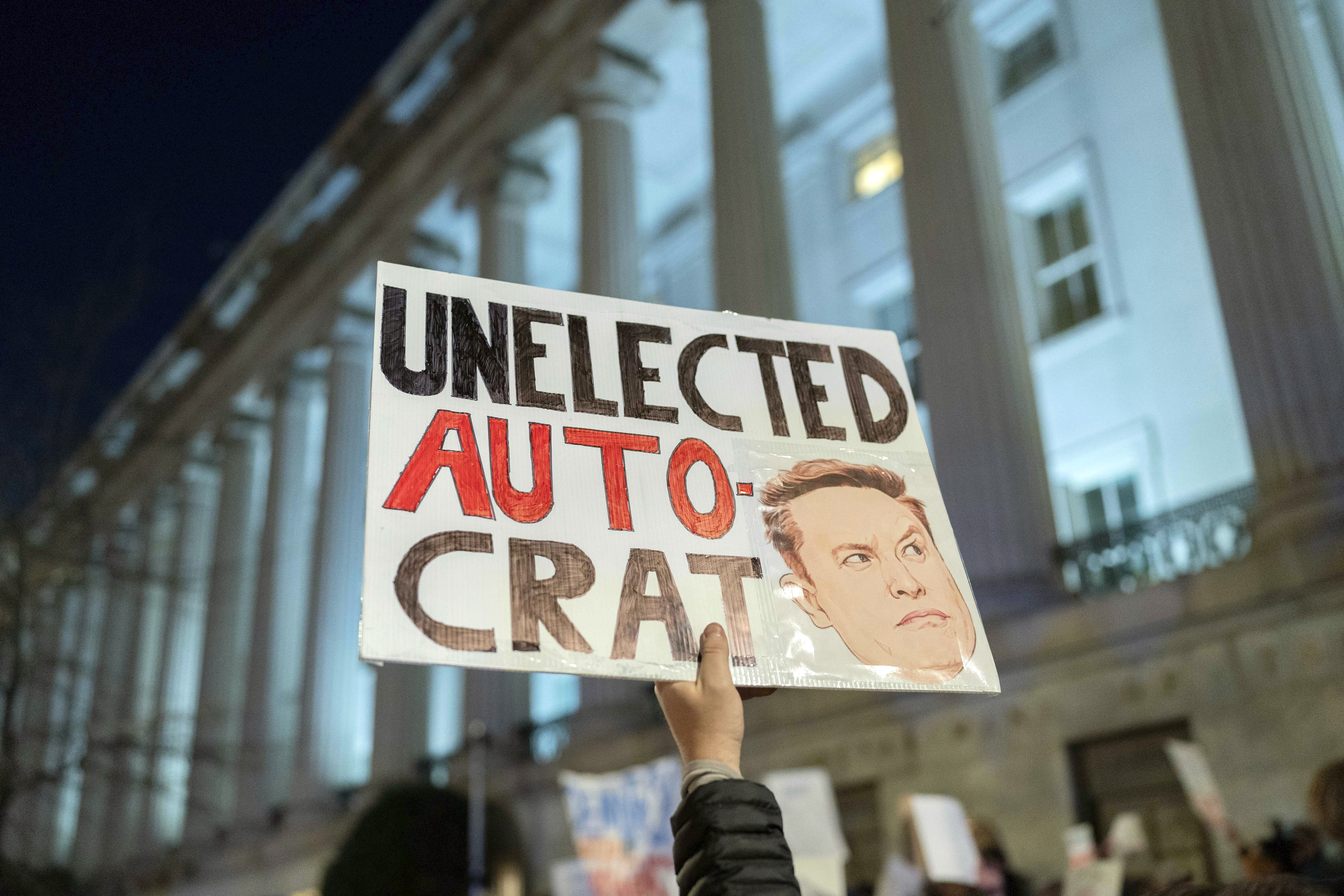 A protester holds up a sign with Elon Musk's face and the words, "unelected autocrat"