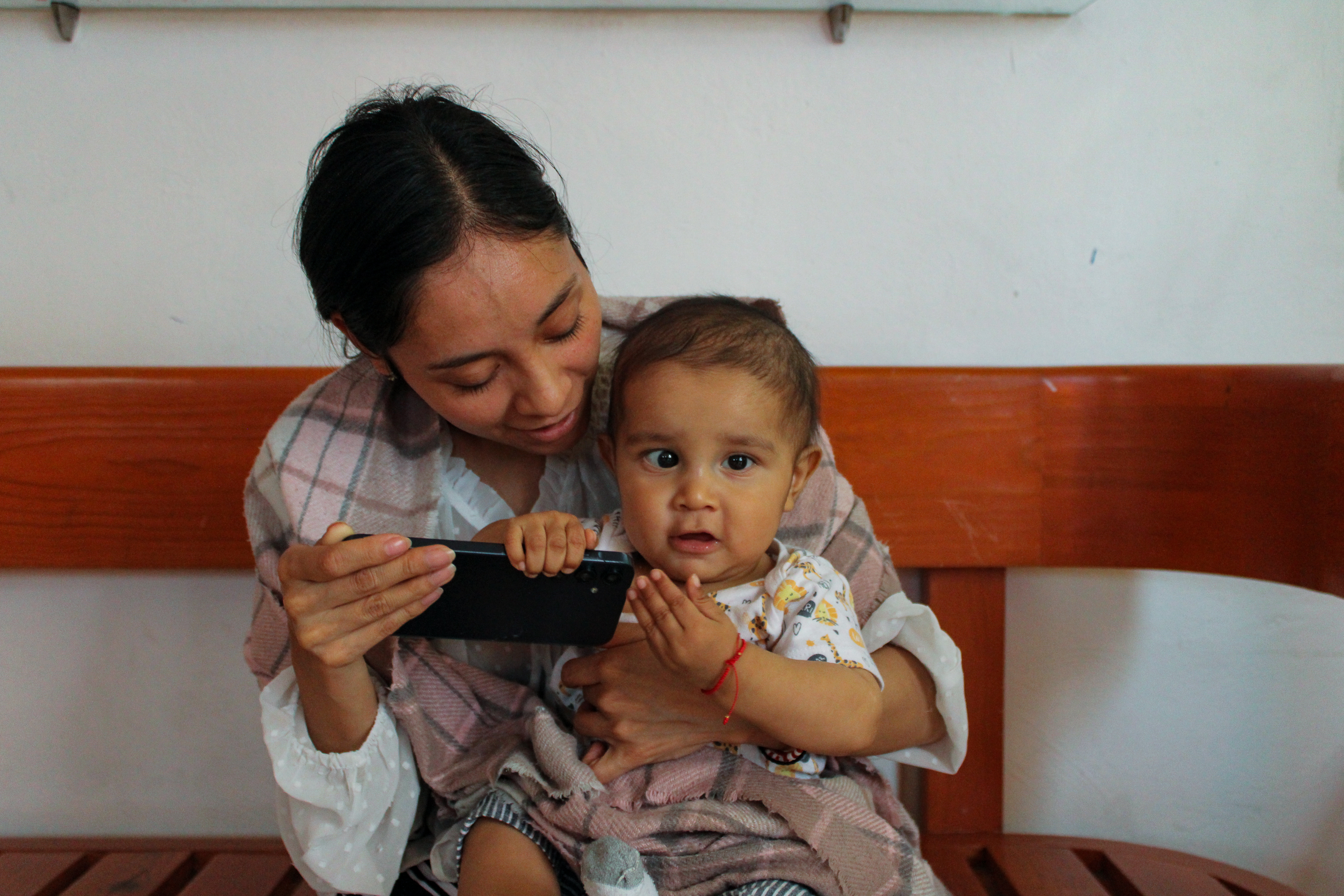 Luisa sits with Juan de Jesús on her knee in the lobby, playing his favourite nursery rhyme, La lechuza (“The Little Owl”), on her phone as they wait to be called_-1773831288