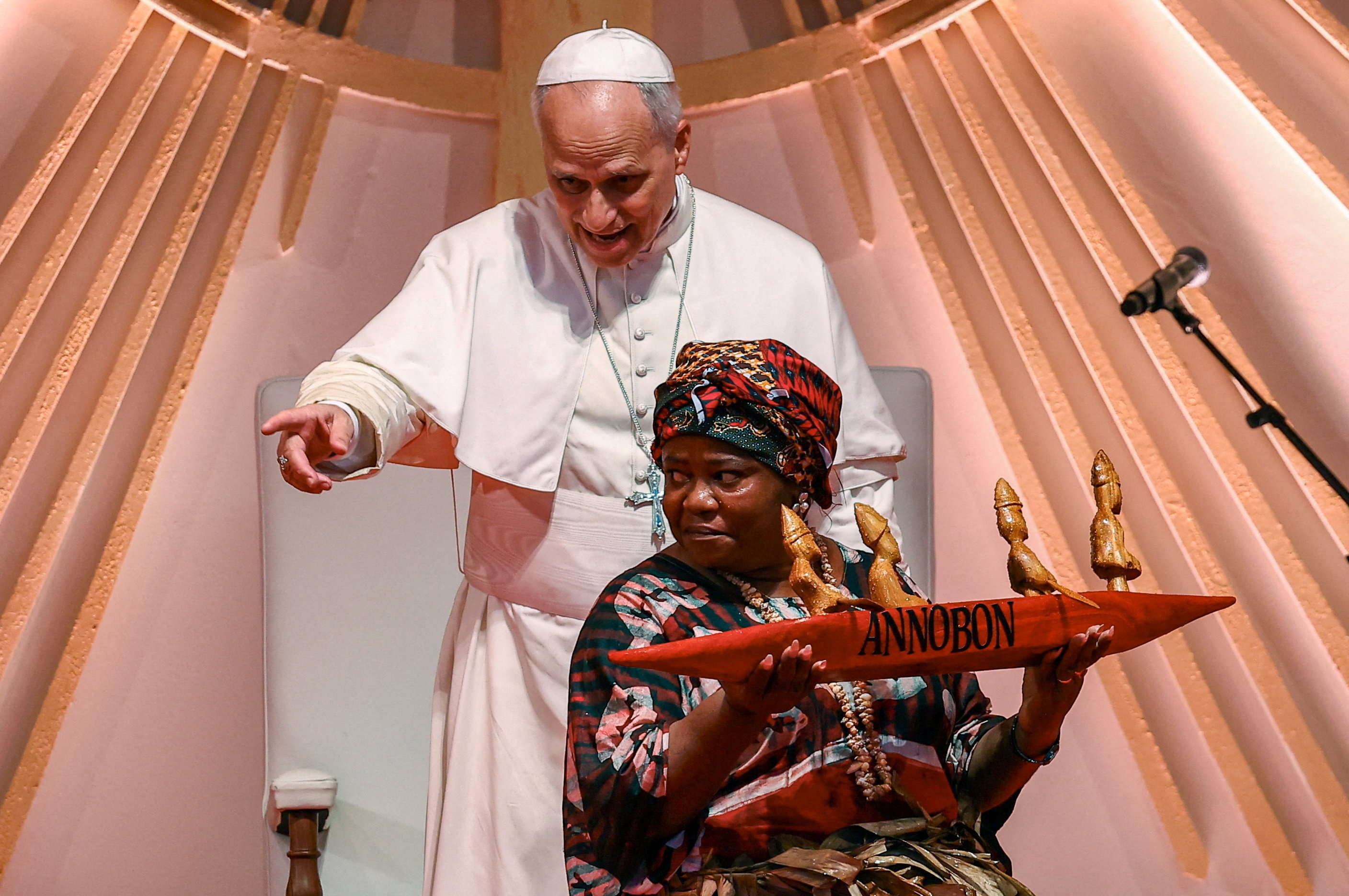 A woman carries a gift for Pope Leo as he points whilst standing behind her