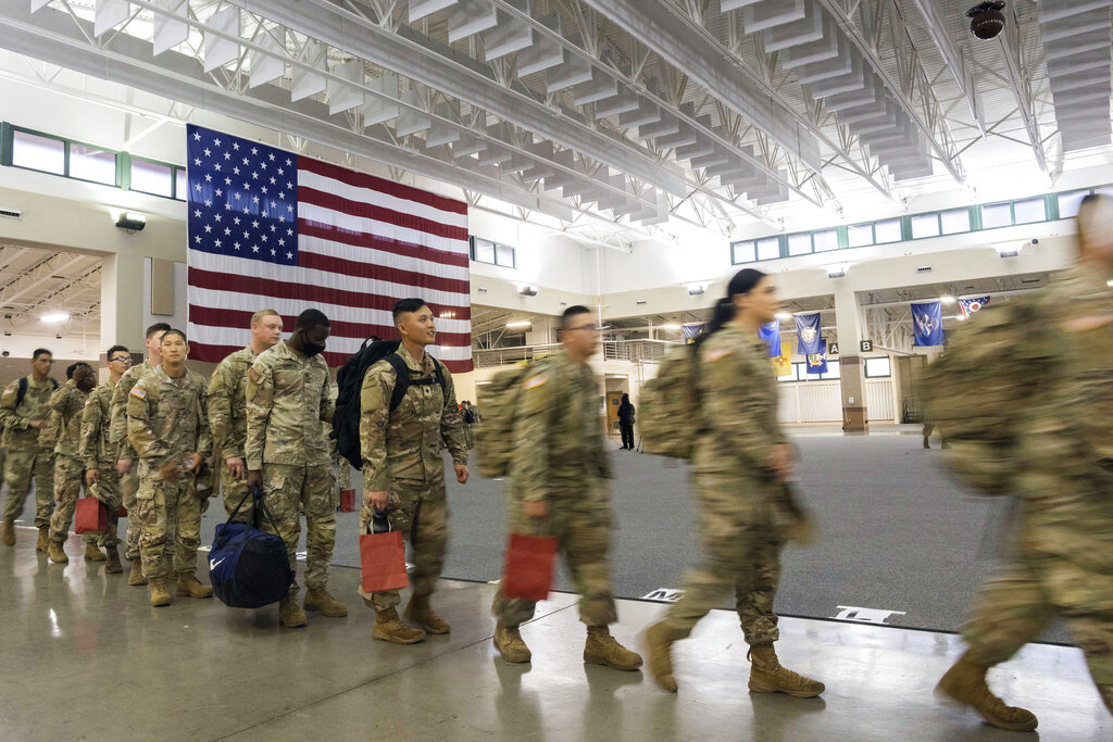 Soldiers wait to board a plane with a US flag in the background