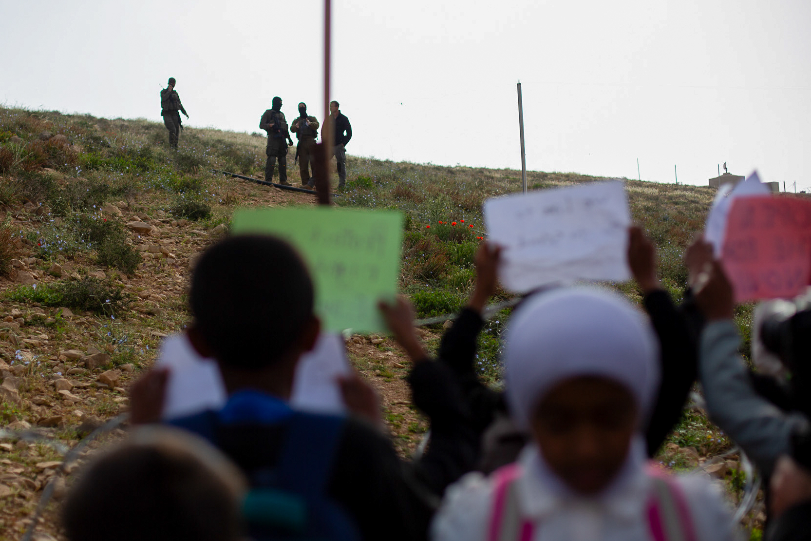 Kids hold up signs as Israeli soldiers watch with settlement security guard